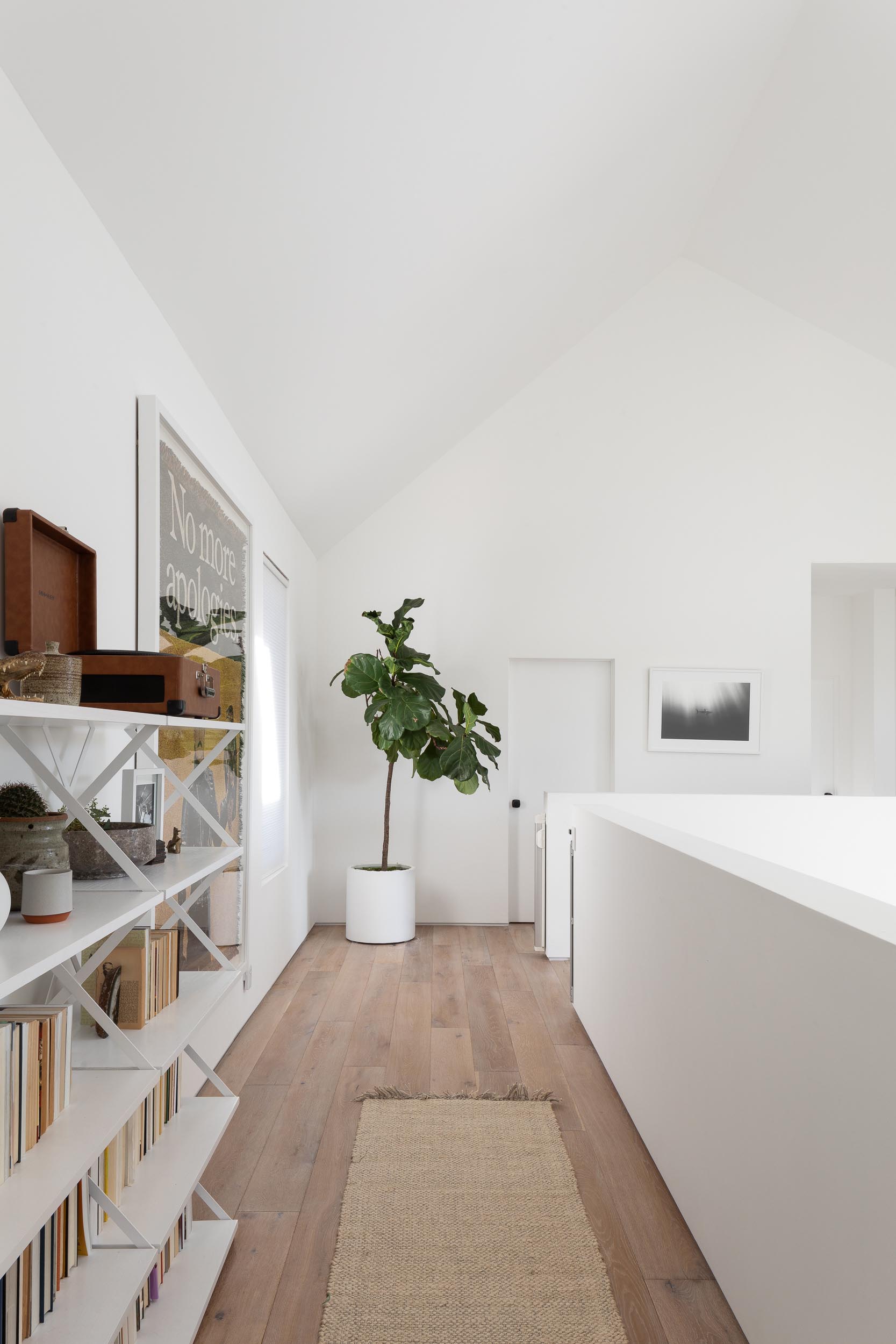 At the top of the stairs in this modern home, there's an open hallway with wood flooring, a tall plant in the corner, artwork, and shelving.