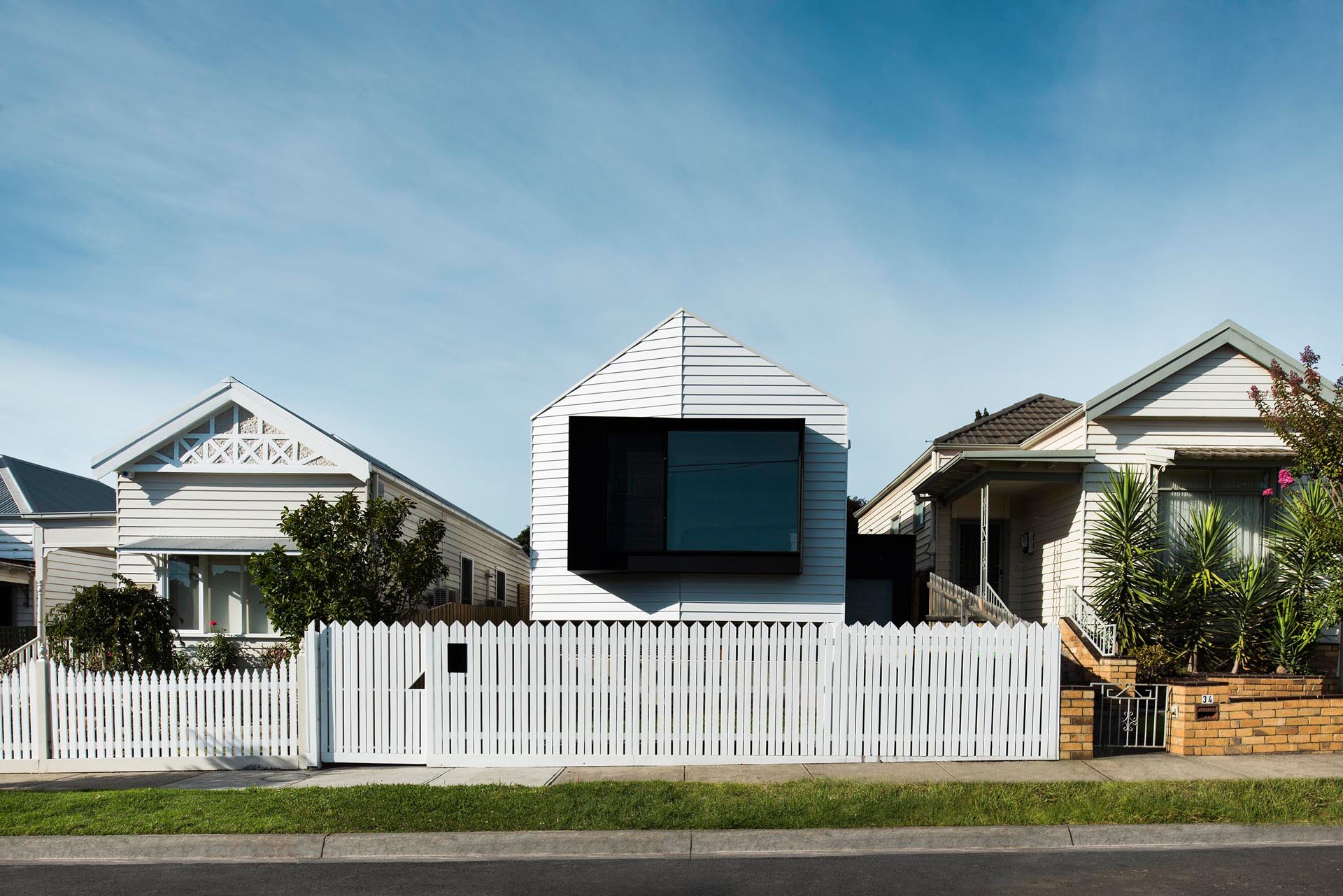 The front of the home has a white exterior which matches the white picket fence, while a large black-framed window protrudes away from the house.