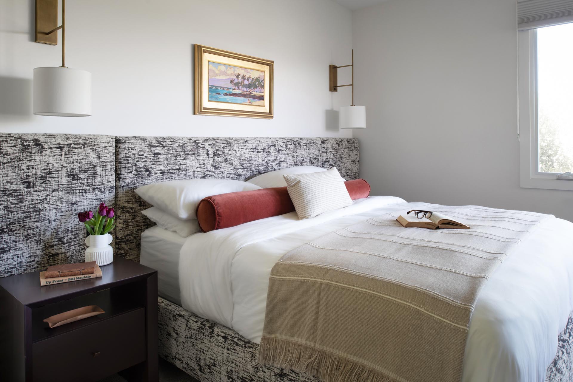 In this primary bedroom, a custom wall to wall headboard in Kelly Wearstler fabric was designed with integrated light switches as well as outlets. The fabric used for the headboard also carries on to the bed frame, while wall lights act as bedside tables.