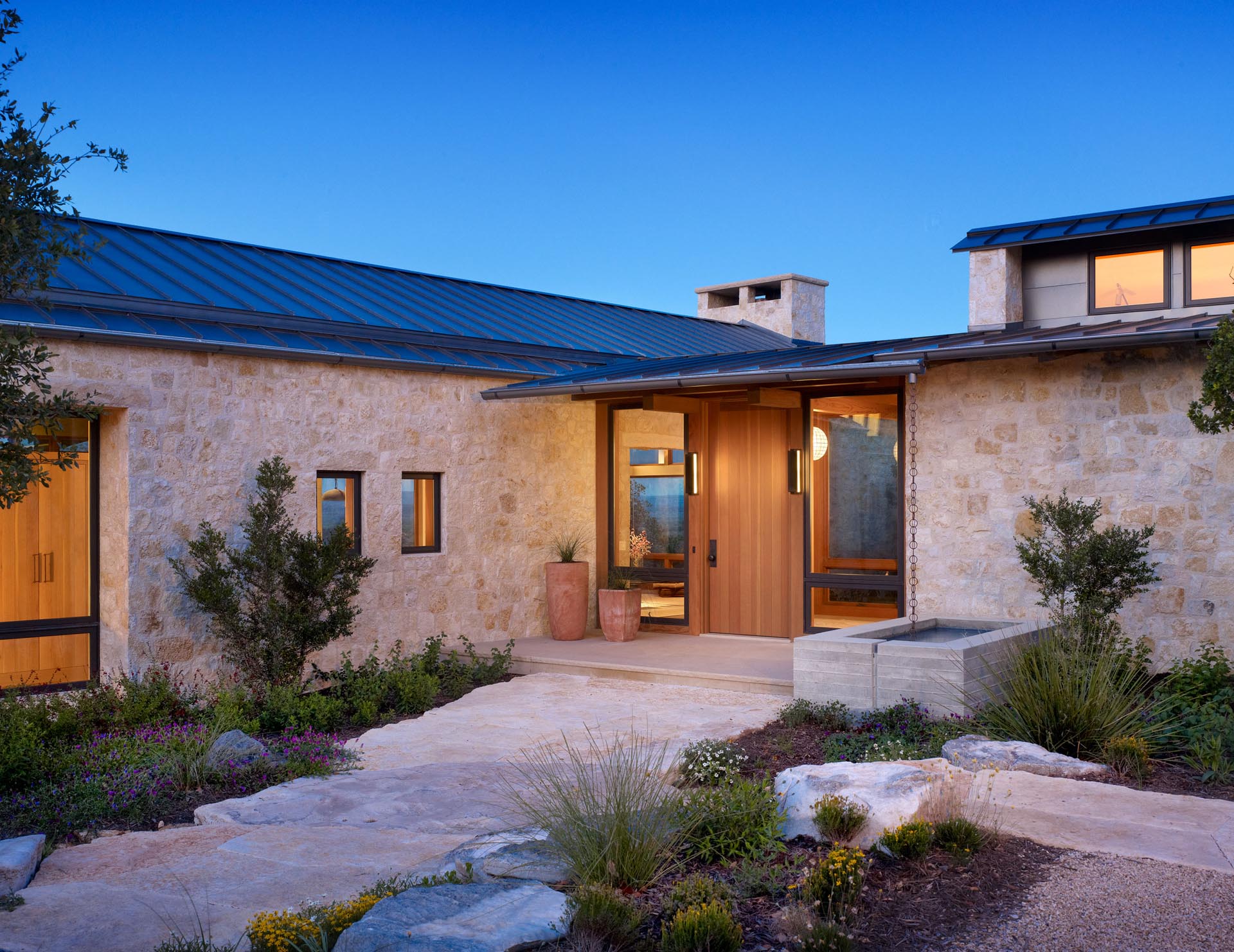 This contemporary home includes limestone walls, a metal roof, black window frames, and a landscaped path to the wood front door.