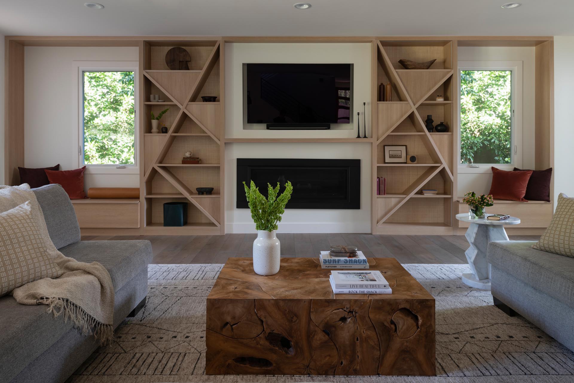 A modern living room with a custom designed wall that includes a recessed TV, a fireplace, wood shelving, and a pair of window seats.