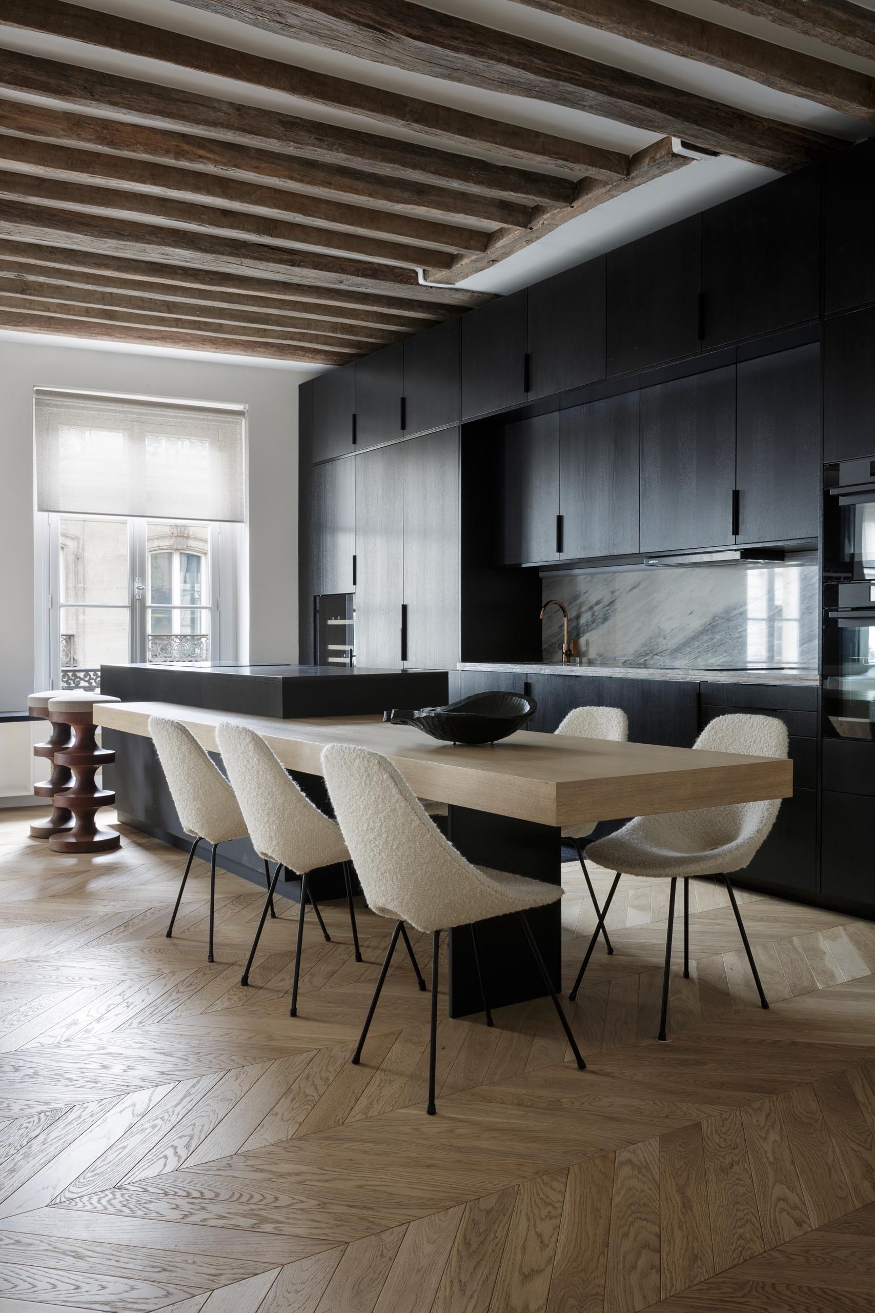 A modern matte black kitchen with a black appliances, exposed wood beams, and an island with a built-in dining table.