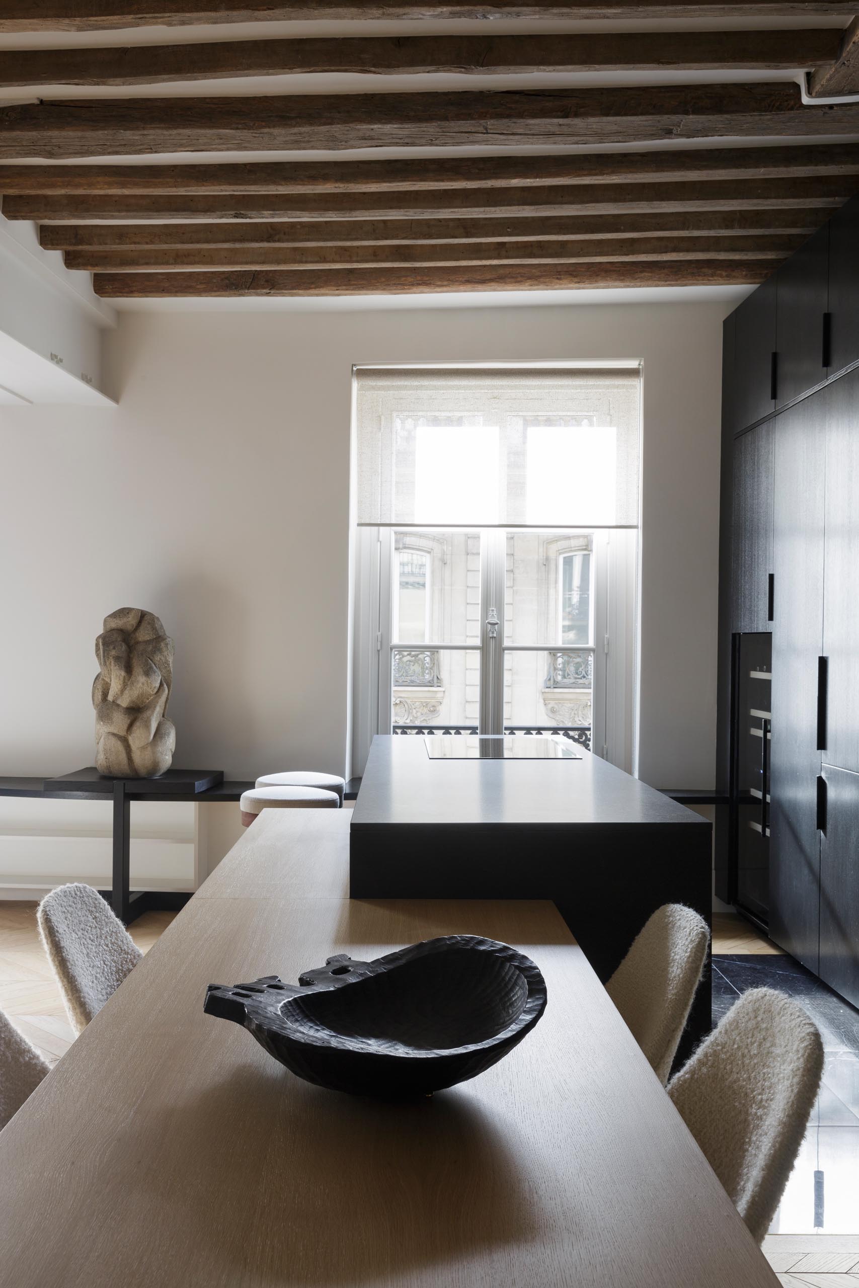 A modern matte black kitchen with a black appliances, exposed wood beams, and an island with a built-in dining table.