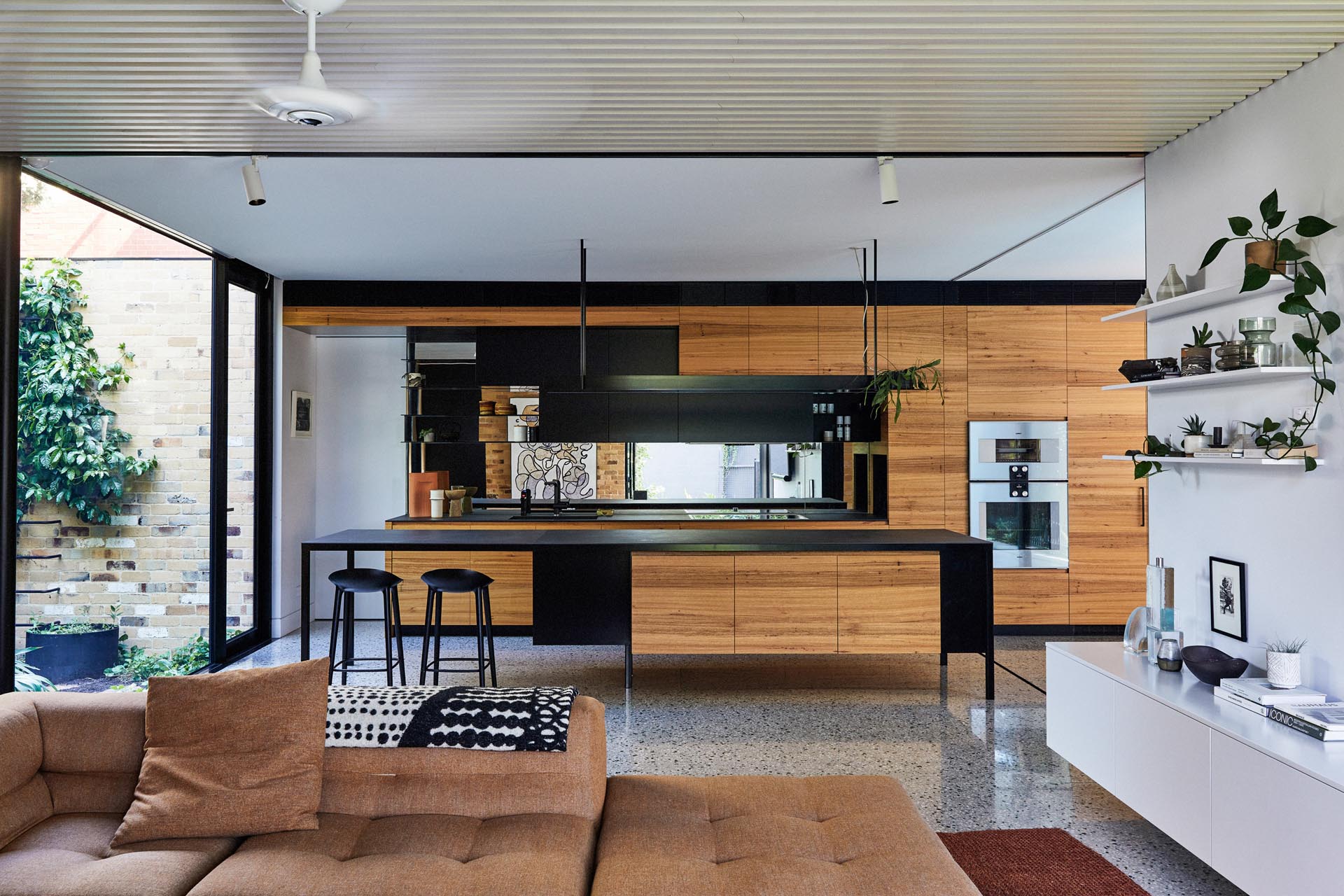 In this modern kitchen, hardware free cabinets line the wall, while the black island and shelving creates a contrasting element, and the mirrored backsplash reflects the living room.