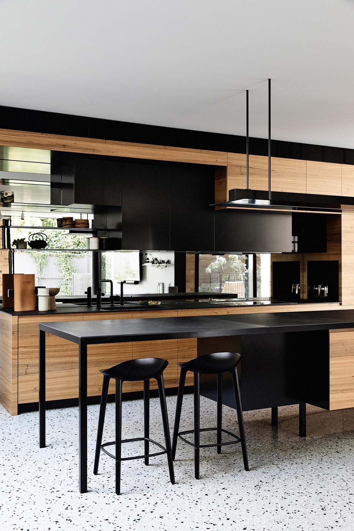 In this modern kitchen, hardware free cabinets line the wall, while the black island and shelving creates a contrasting element, and the mirrored backsplash reflects the living room.