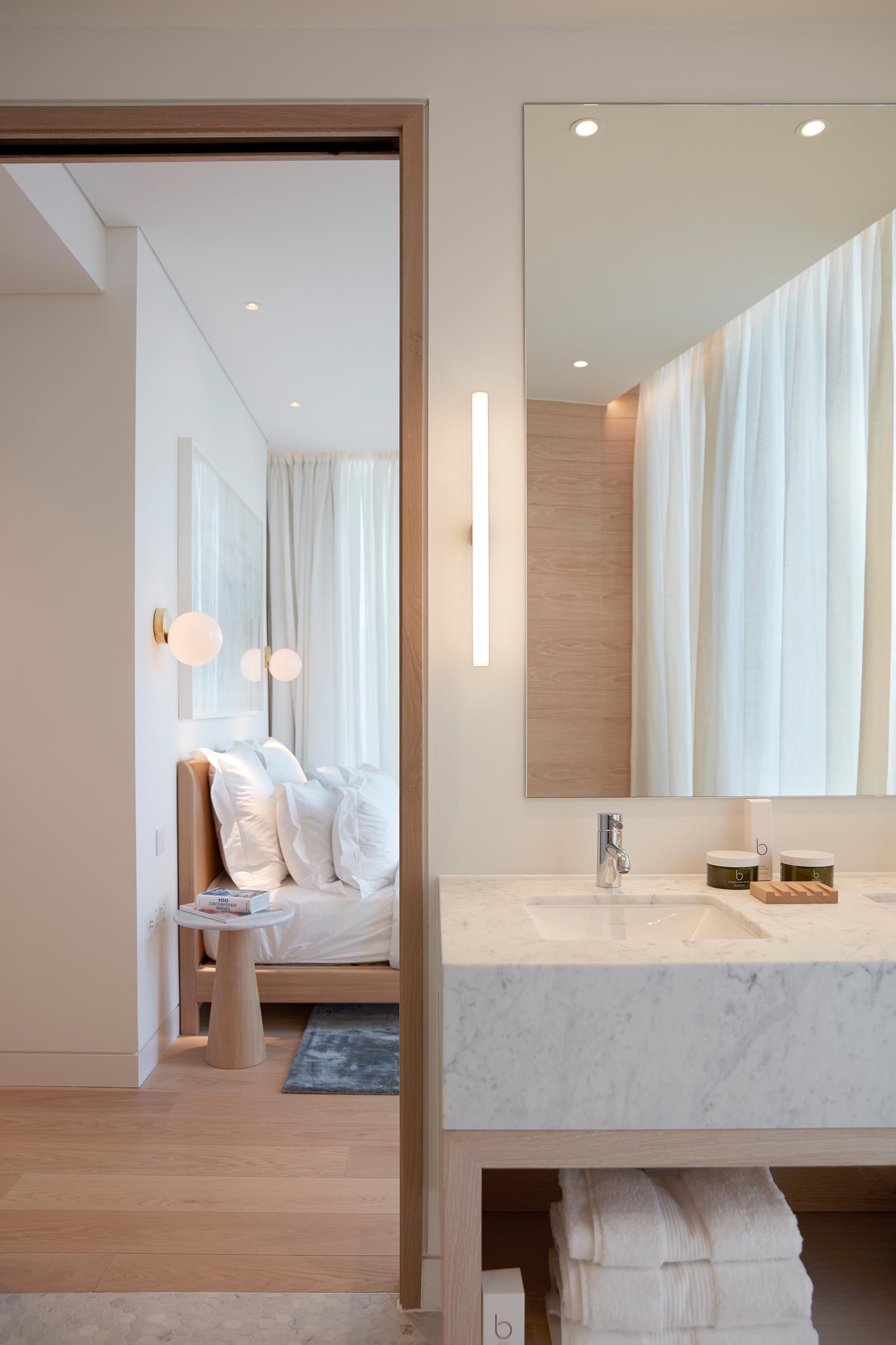 In an modern bathroom, light wood has been used for the open vanity shelving, as well as the accent wall that's installed behind the freestanding white bathtub, while the vanity countertop complements the walk-in shower.