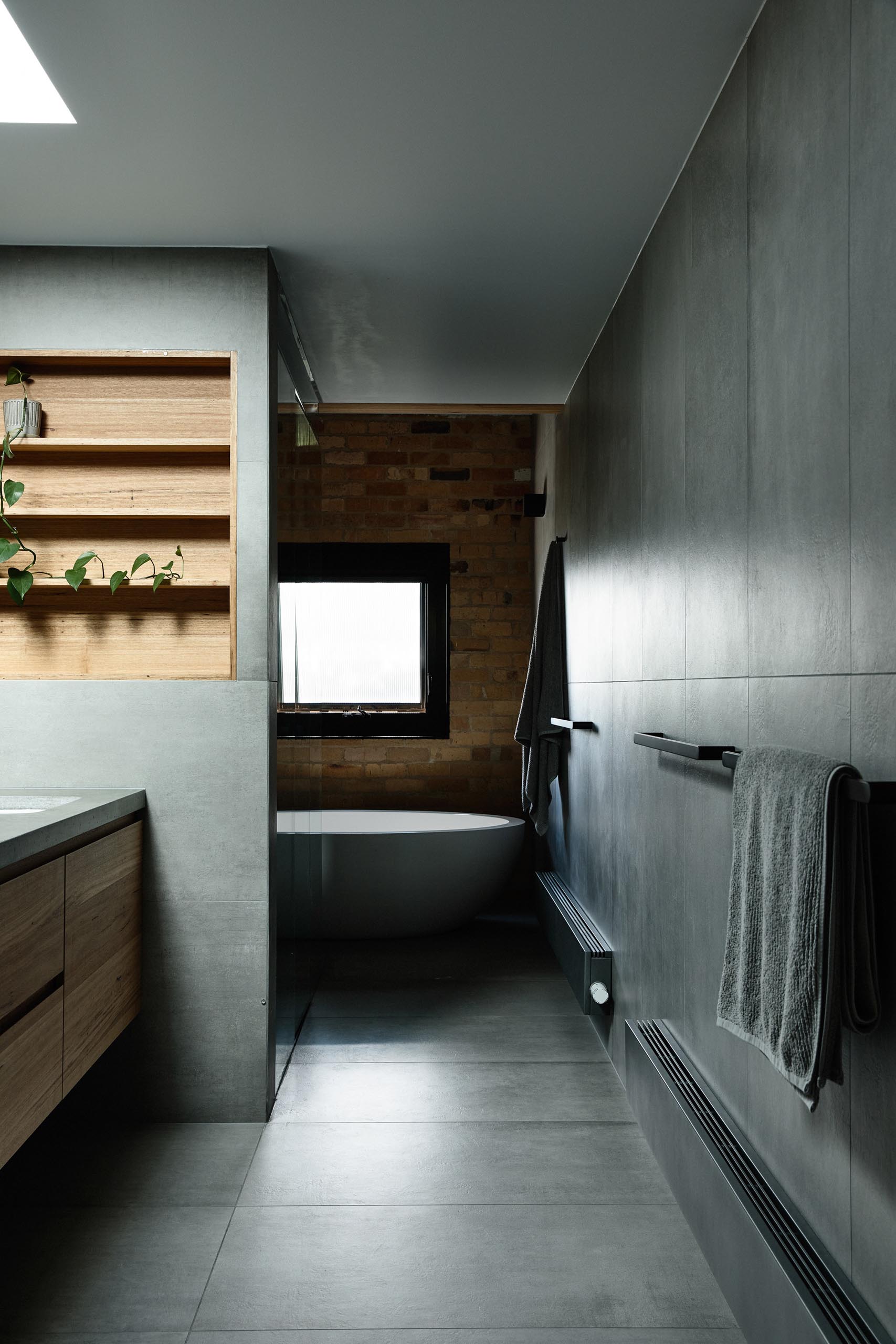 In this modern bathroom, gray walls and floors are accented by a wood-lined shelving niche, small metallic tiles, and a white freestanding bathtub.