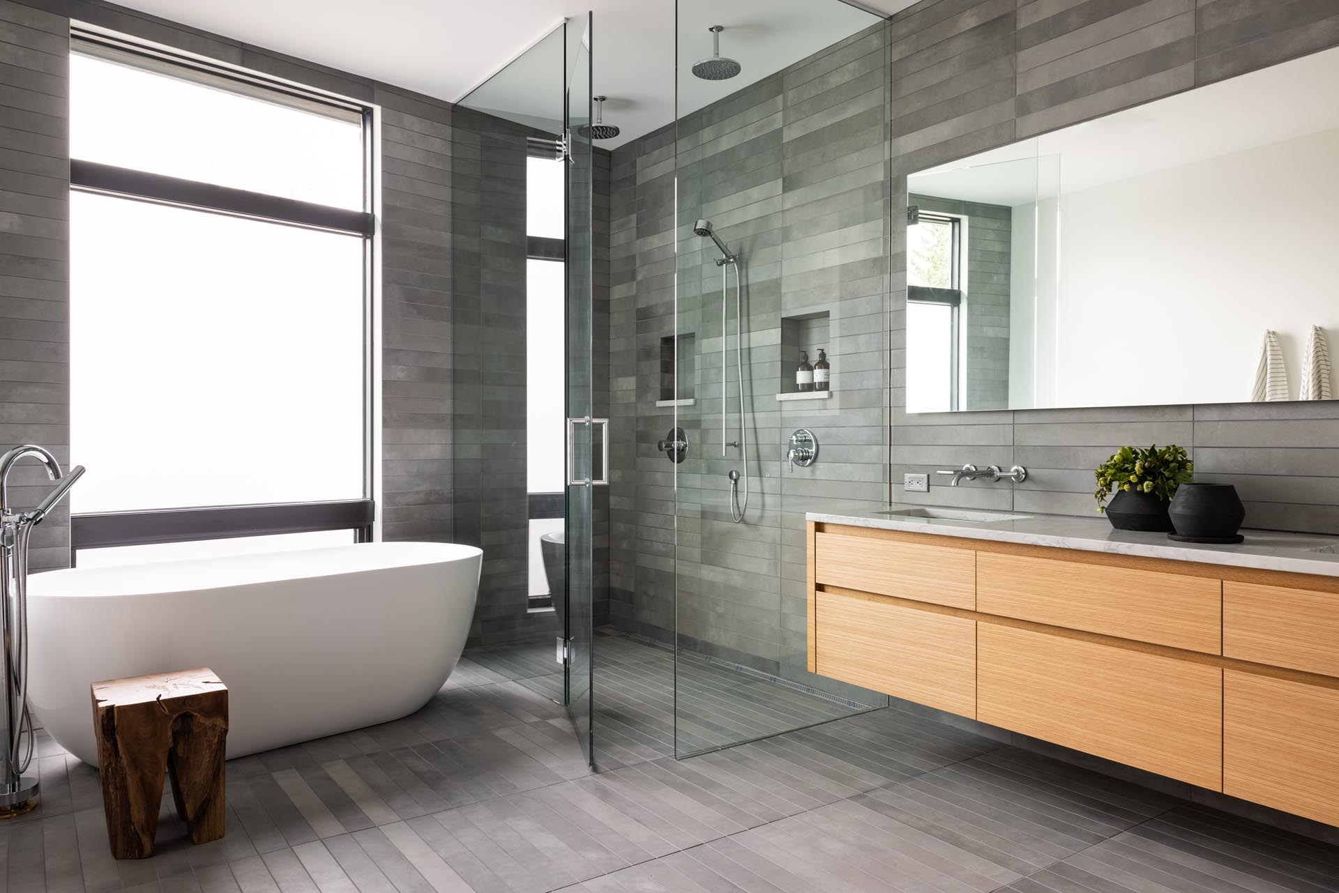 In this modern bathroom, grey tiles cover the walls and floor, while a floating wood vanity adds a natural element, and a white freestanding tub is positioned by the glass-enclosed shower.