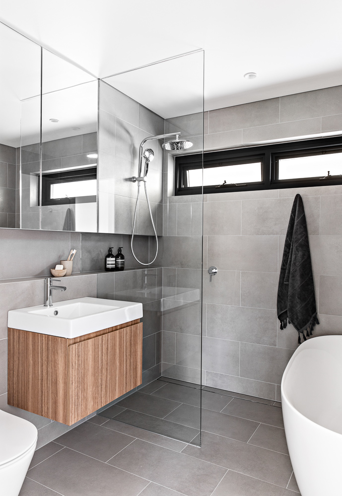 A modern bathroom with large gray tiles that cover the walls and floor, a floating wood vanity, and a shower niche.