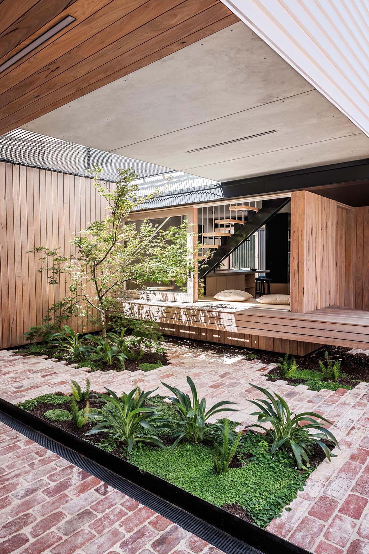 Connecting the interior spaces with the carport of this modern home, is a small courtyard with bricks surrounding small pockets of greenery. In the open space above, there's a net for relaxing in the sun.