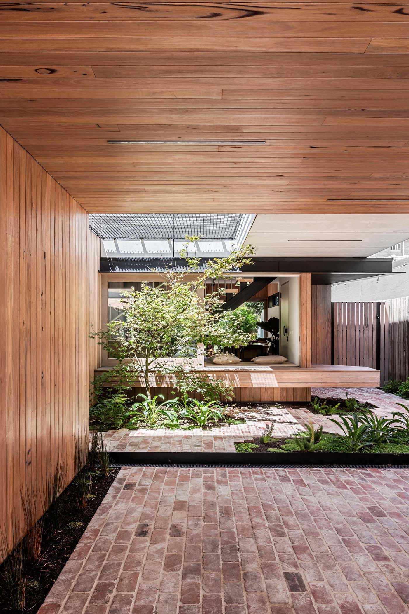 Connecting the interior spaces with the carport of this modern home, is a small courtyard with bricks surrounding small pockets of greenery. In the open space above, there's a net for relaxing in the sun.
