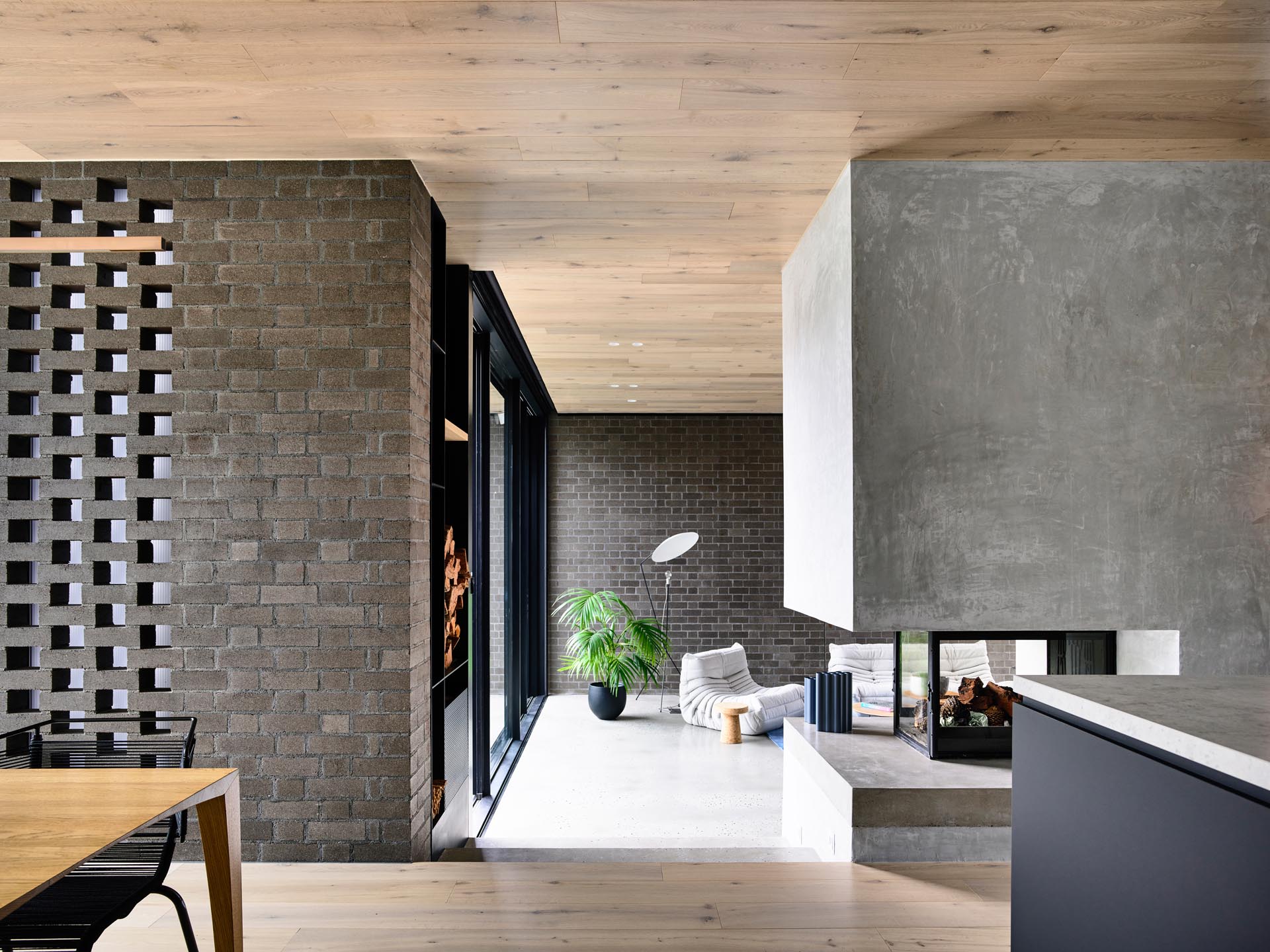 This modern interior showcases dark brick walls, as well as a wood ceiling, and a see-through fireplace that can be enjoyed from both the kitchen and the living room.