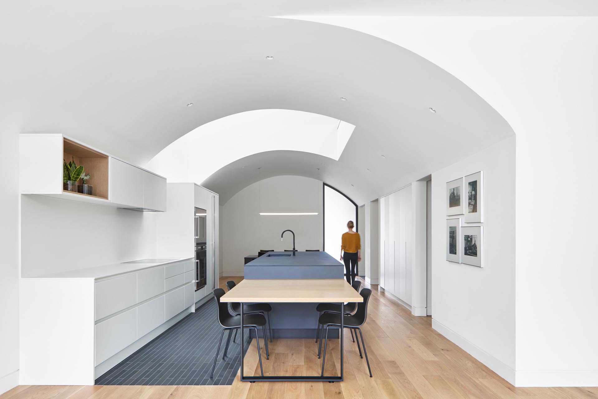 This kitchen, which has white minimalist cabinets that line the wall, and a matte blue kitchen island, also includes a more casual dining area by the living room.