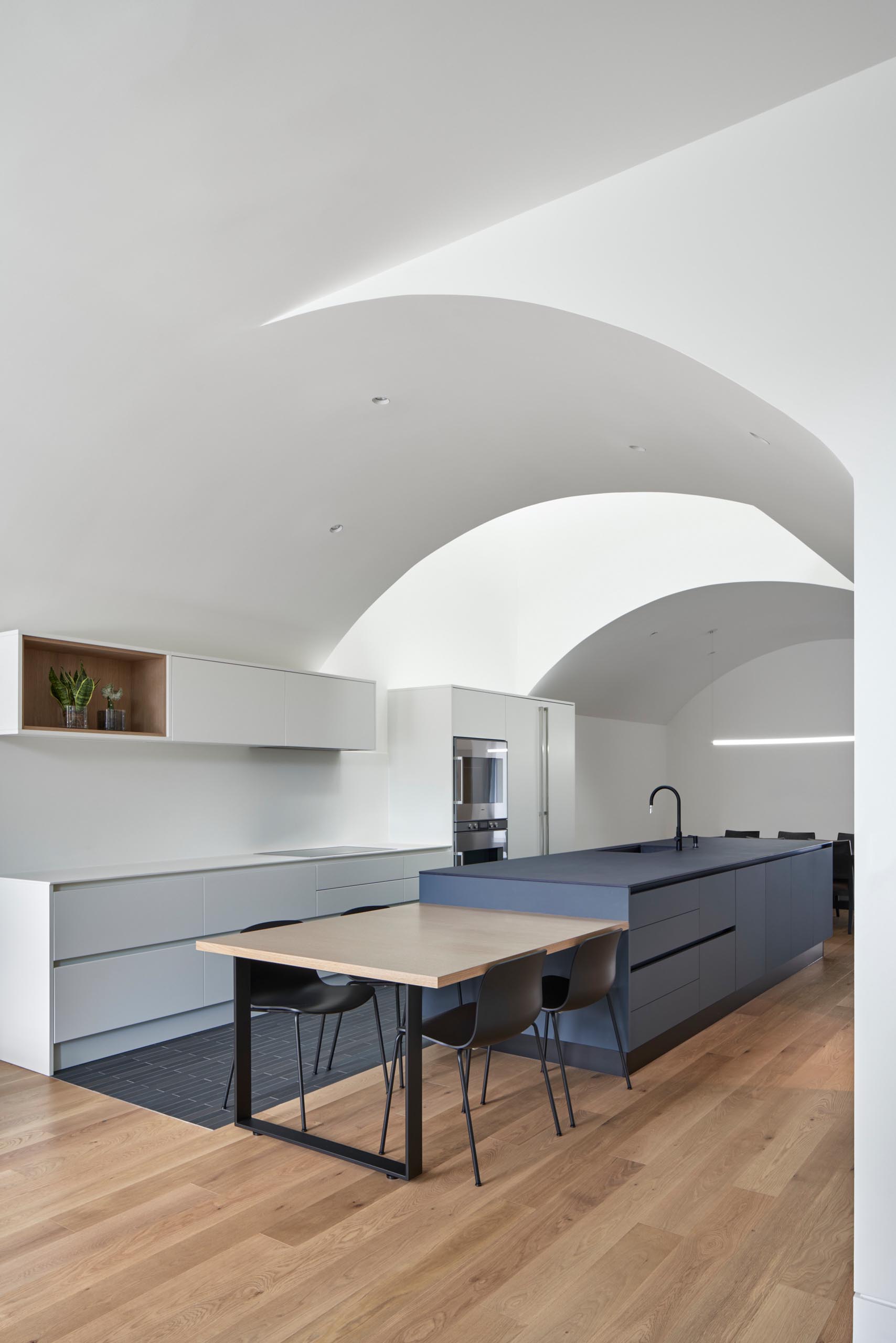 This kitchen, which has white minimalist cabinets that line the wall, and a matte blue kitchen island, also includes a more casual dining area by the living room.