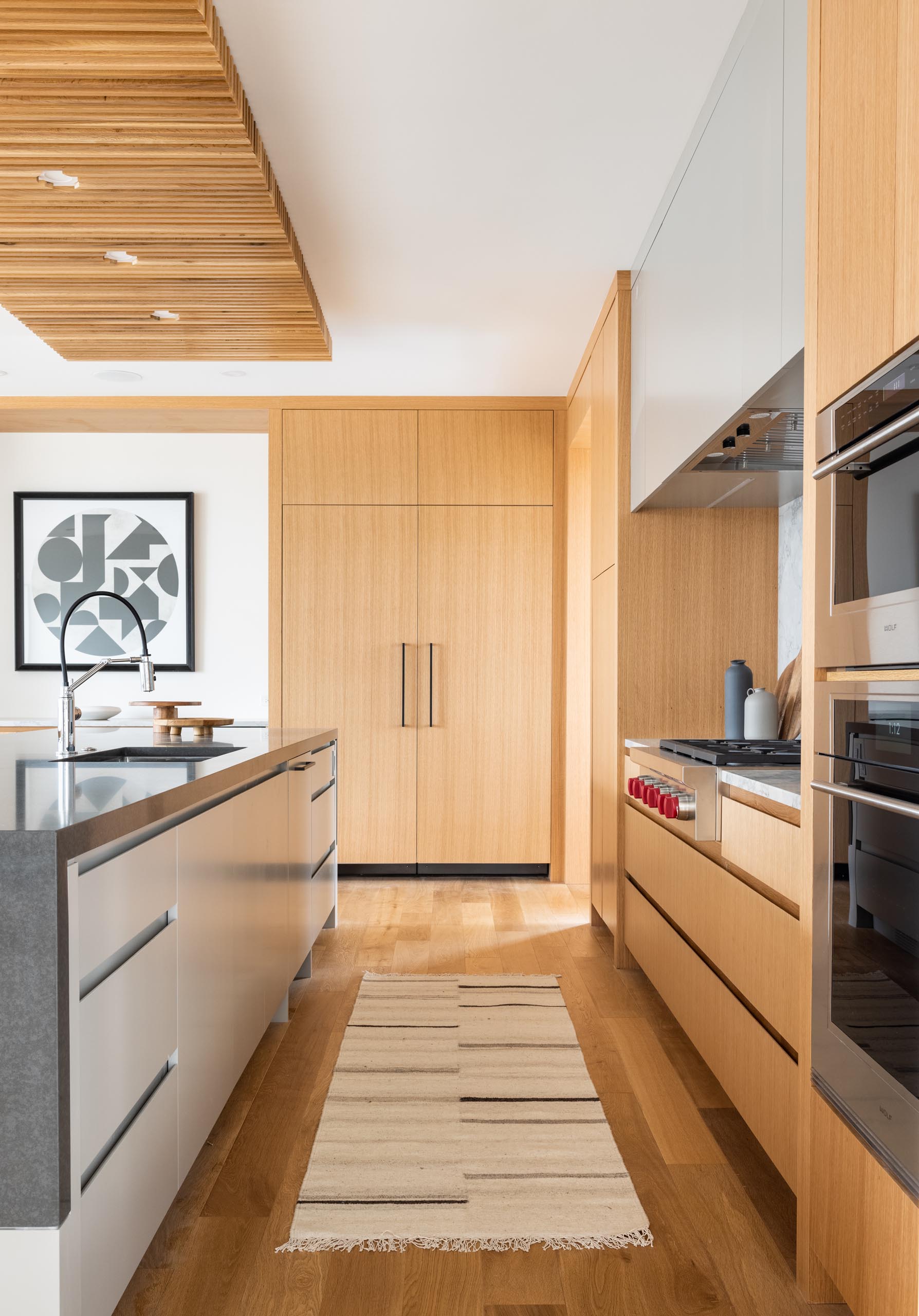 This modern kitchen includes a large island with a wood ceiling accent and light wood cabinets.