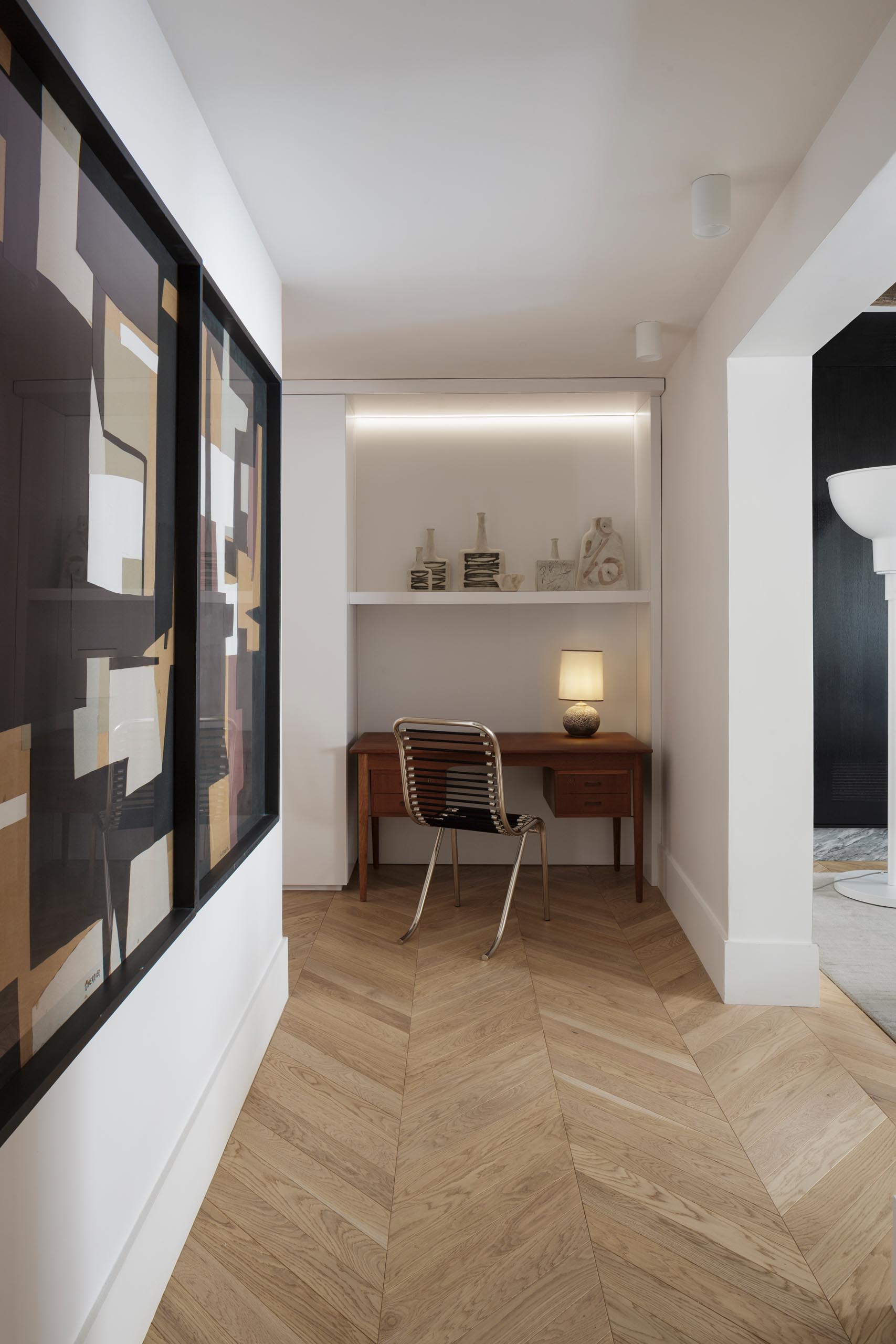 A small home office is located at the end of a hallway, and includes a small wood desk as well as a built-in shelf and hidden lighting.