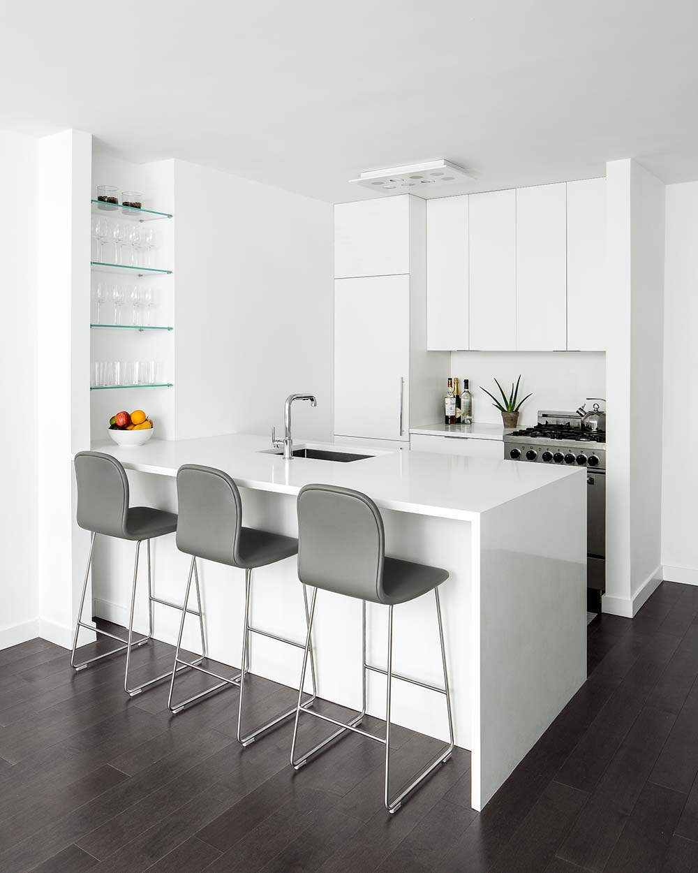 A modern white kitchen with an integrated fridge, a peninsula with a waterfall countertop, and a shelving niche with glass shelves.