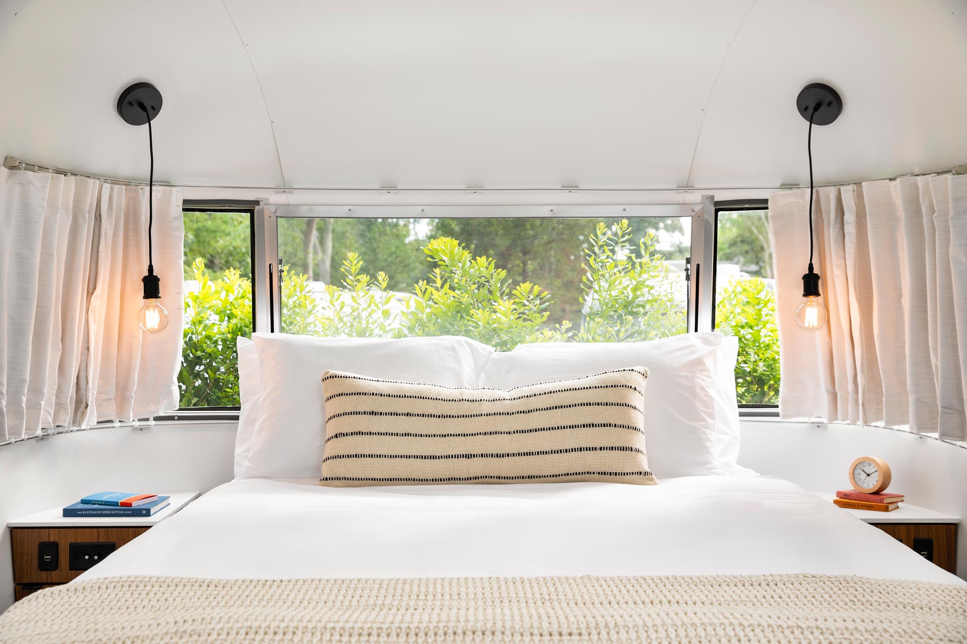 A remodeled Airstream bedroom is furnished with a queen bed, built-in side tables, and a pair of simple pendant lights.