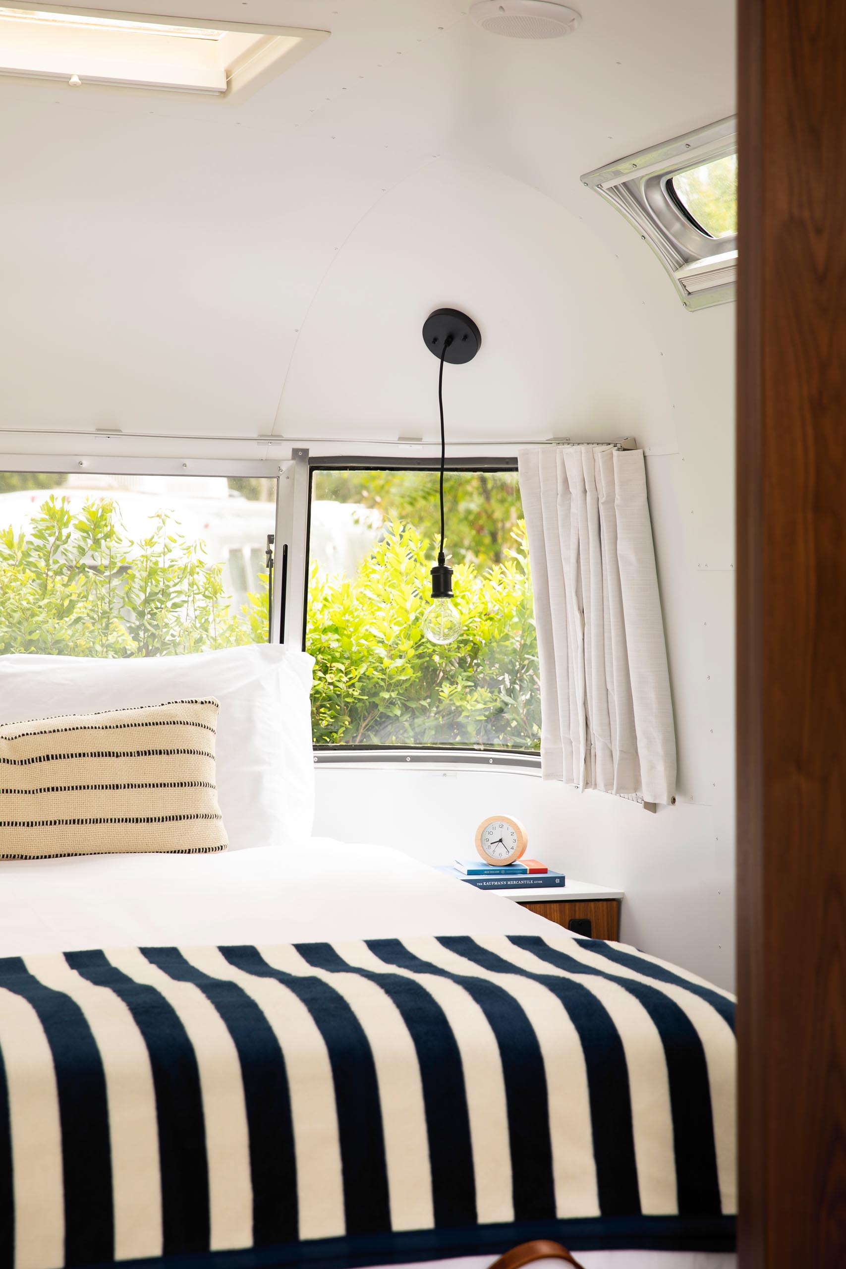 A remodeled Airstream bedroom is furnished with a queen bed, built-in side tables, and a pair of simple pendant lights. A panoramic window fills the small space with natural light and views of the gardens, while at night the curtains can be closed for a more cozy and private atmosphere.