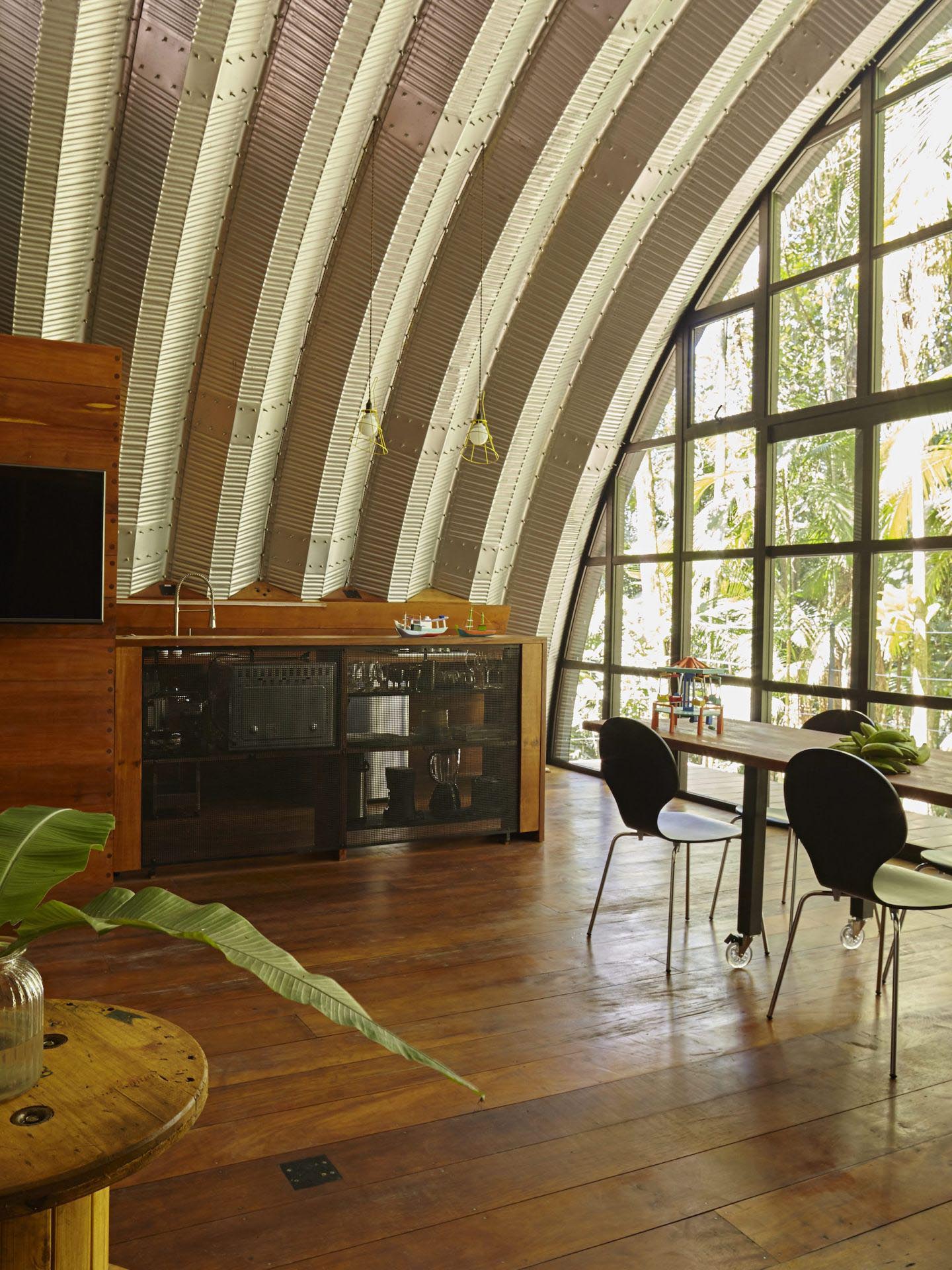 A modern home with an arched metal roof has a small kitchen, and a full wall of windows.