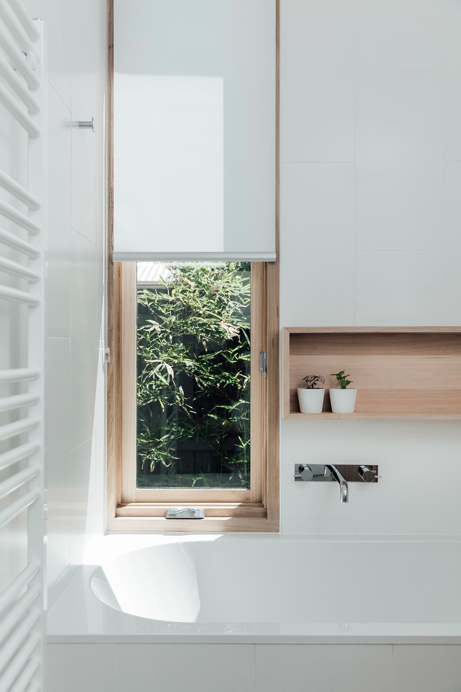 This modern bathroom includes a built-in bathtub with a tall window and wood-lined shelving niche on the adjacent wall. A white heated towel rail almost blends into the surrounding white wall.