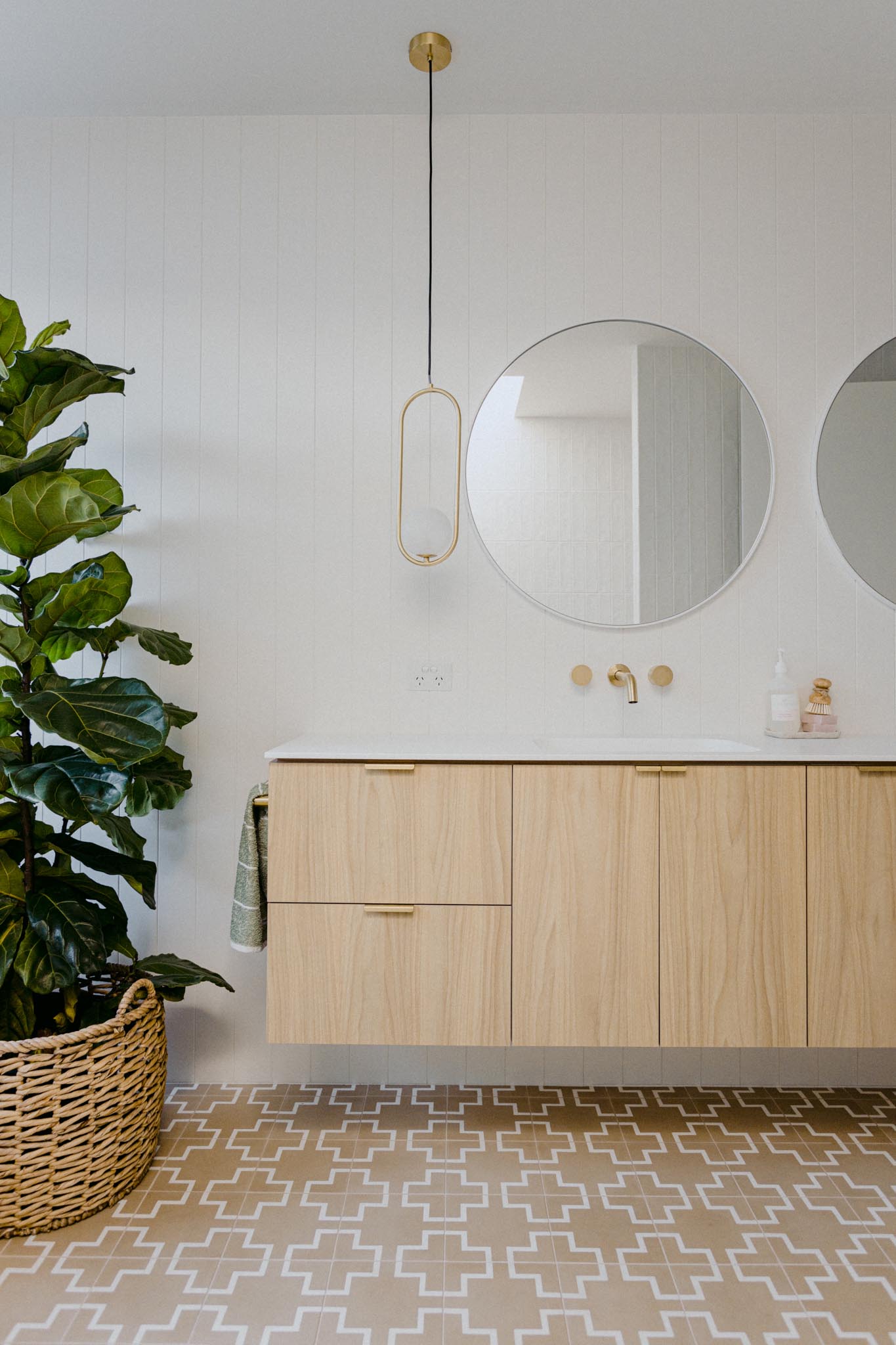 This modern bathroom includes a floating wood vanity with white countertop, round mirrors, white wall tiles, a minimalist pendant light, and patterned tile floor.