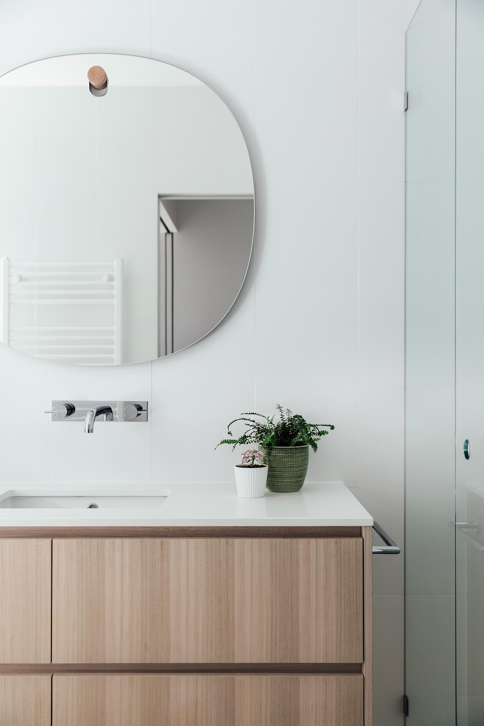 In this modern bathroom, while tiles cover the walls, while a rounded frameless mirror hangs from a simple wood hook, which also matches the wood vanity.