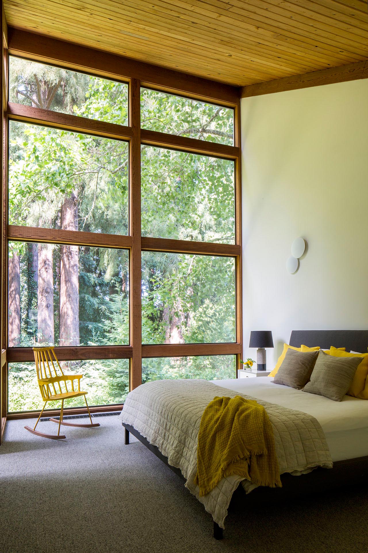 A modern bedroom with high ceilings and a wall of wood-framed windows.