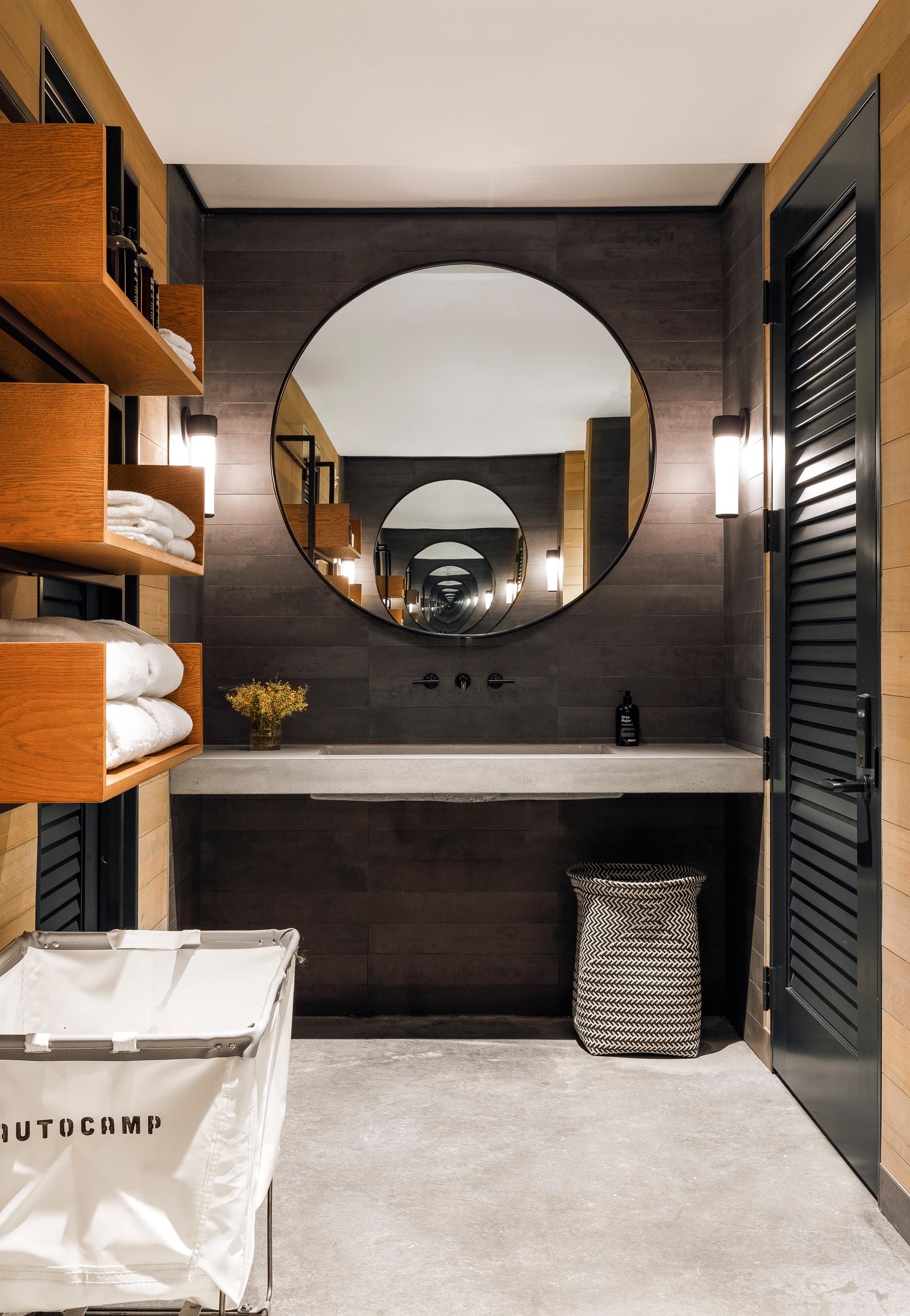 A modern bathroom with dark wall tiles that complement the black frame of the round mirror, as well as the faucet and nearby door.