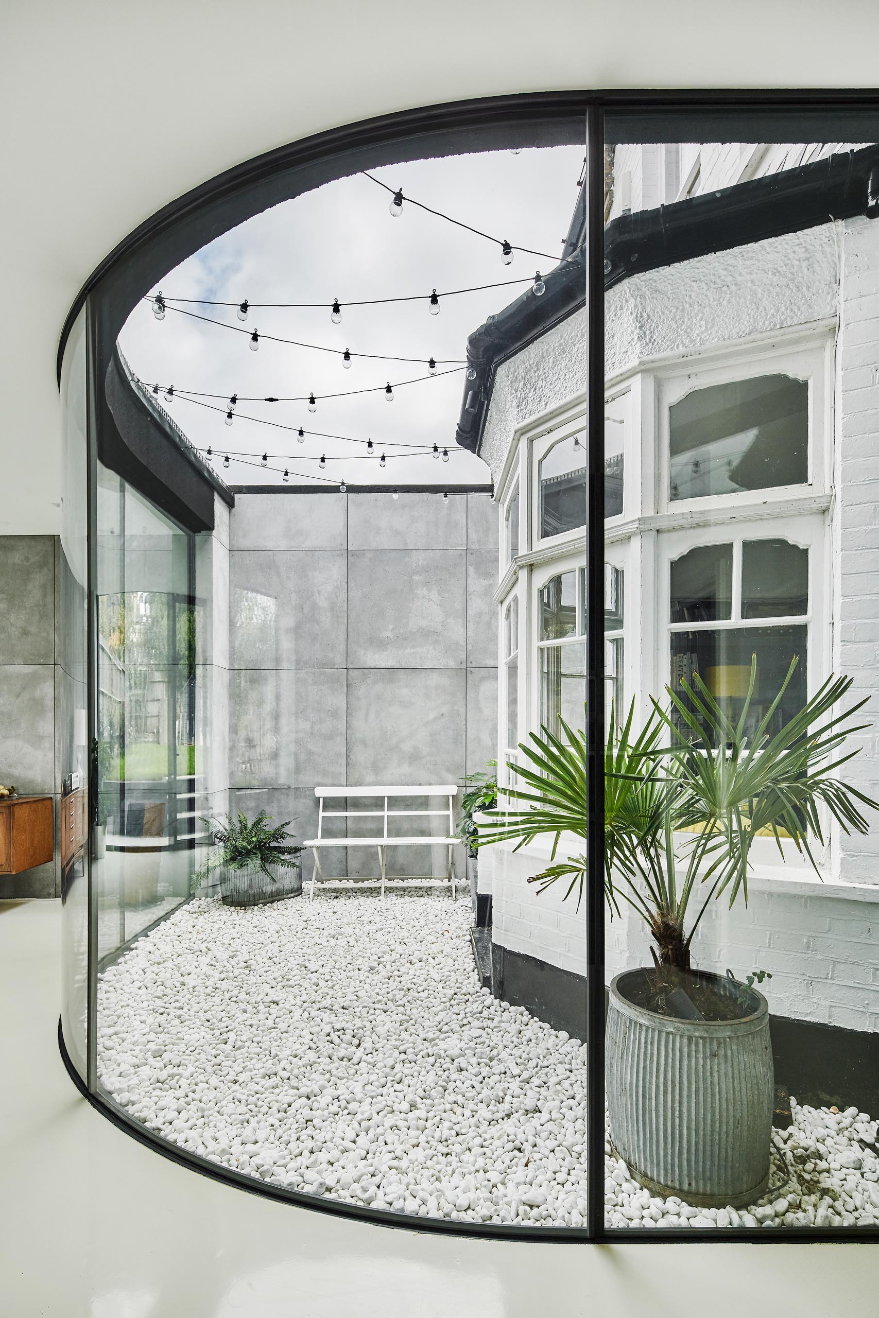 The small open-air courtyard between the bay window and the frameless window can be accessed from within the original home, and includes a white pebble ground cover, some planters, and string lights.