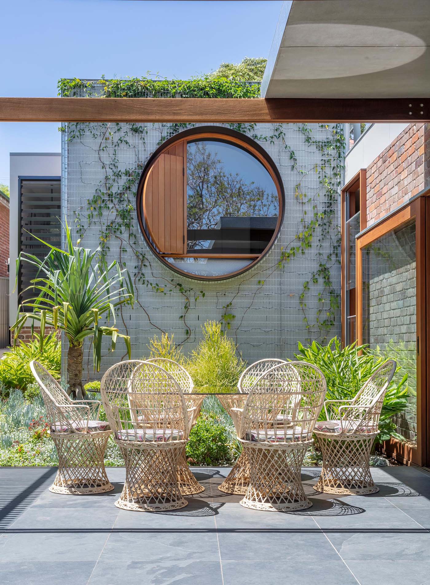 This round outdoor dining table is surrounded by chairs with curved edges, complementing the shape of the nearby circular window.