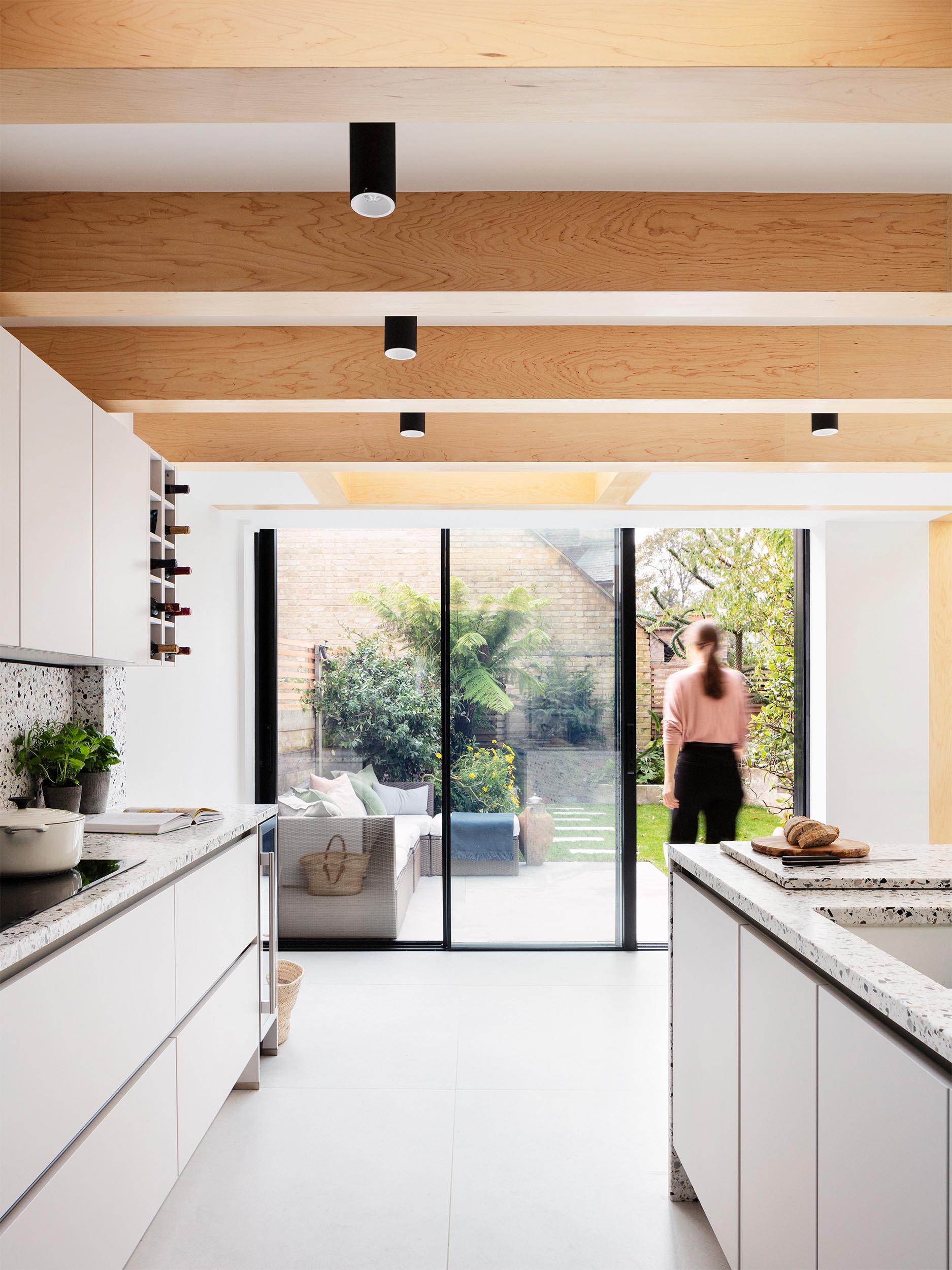 The warmth of the wood beams is mirrored in the neutral tones of the terrazzo that covers the kitchen island and countertops below, and in the muted green paintwork of cabinetry.