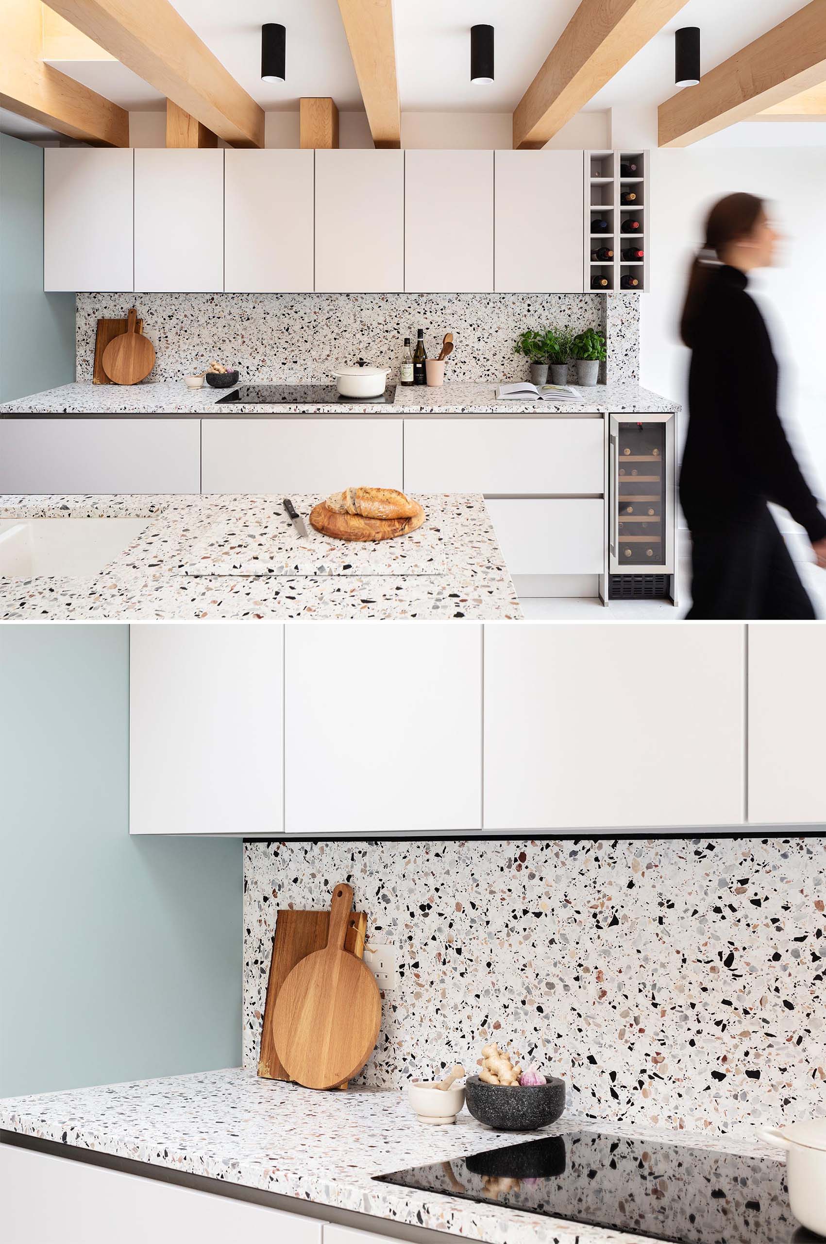 The warmth of the wood beams is mirrored in the neutral tones of the terrazzo that covers the kitchen island and countertops below, and in the muted green paintwork of cabinetry.