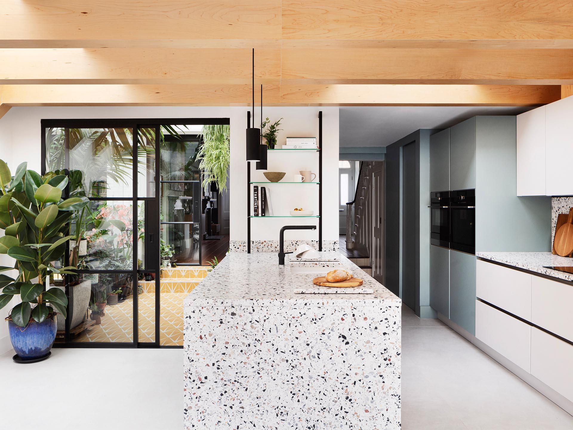 The warmth of the wood beams is mirrored in the neutral tones of the terrazzo that covers the kitchen island and countertops below, and in the muted green paintwork of cabinetry.