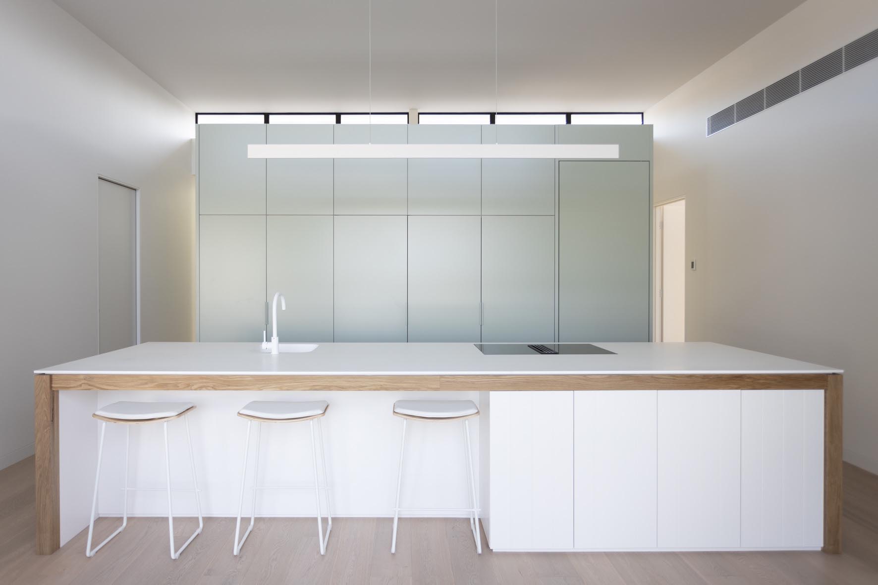 This kitchen has a minimalist design that includes an integrated fridge, and a large white island with wood detailing.