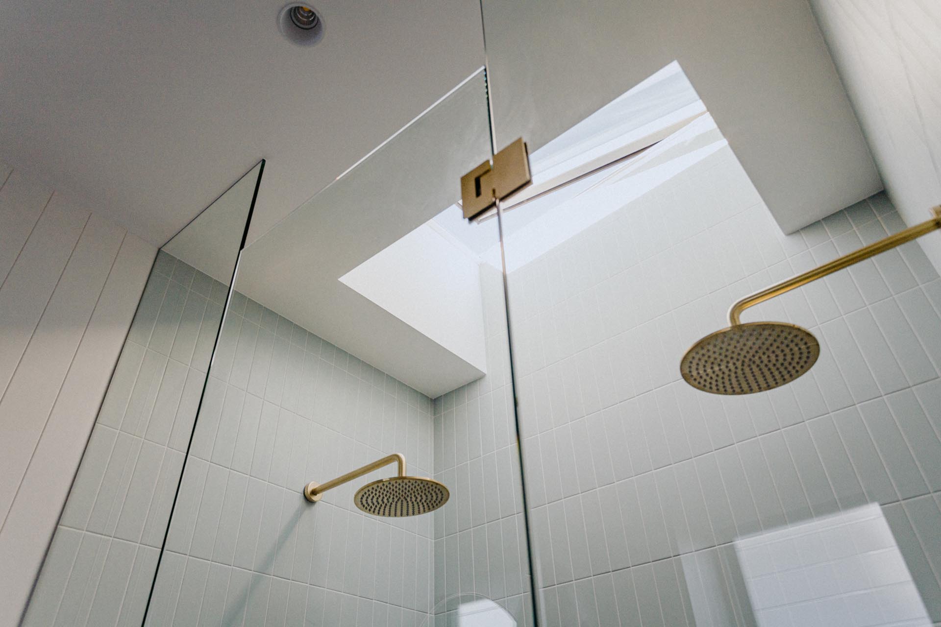 In this modern shower, there's dual shower heads, a frameless glass door, white tiles, and a skylight.