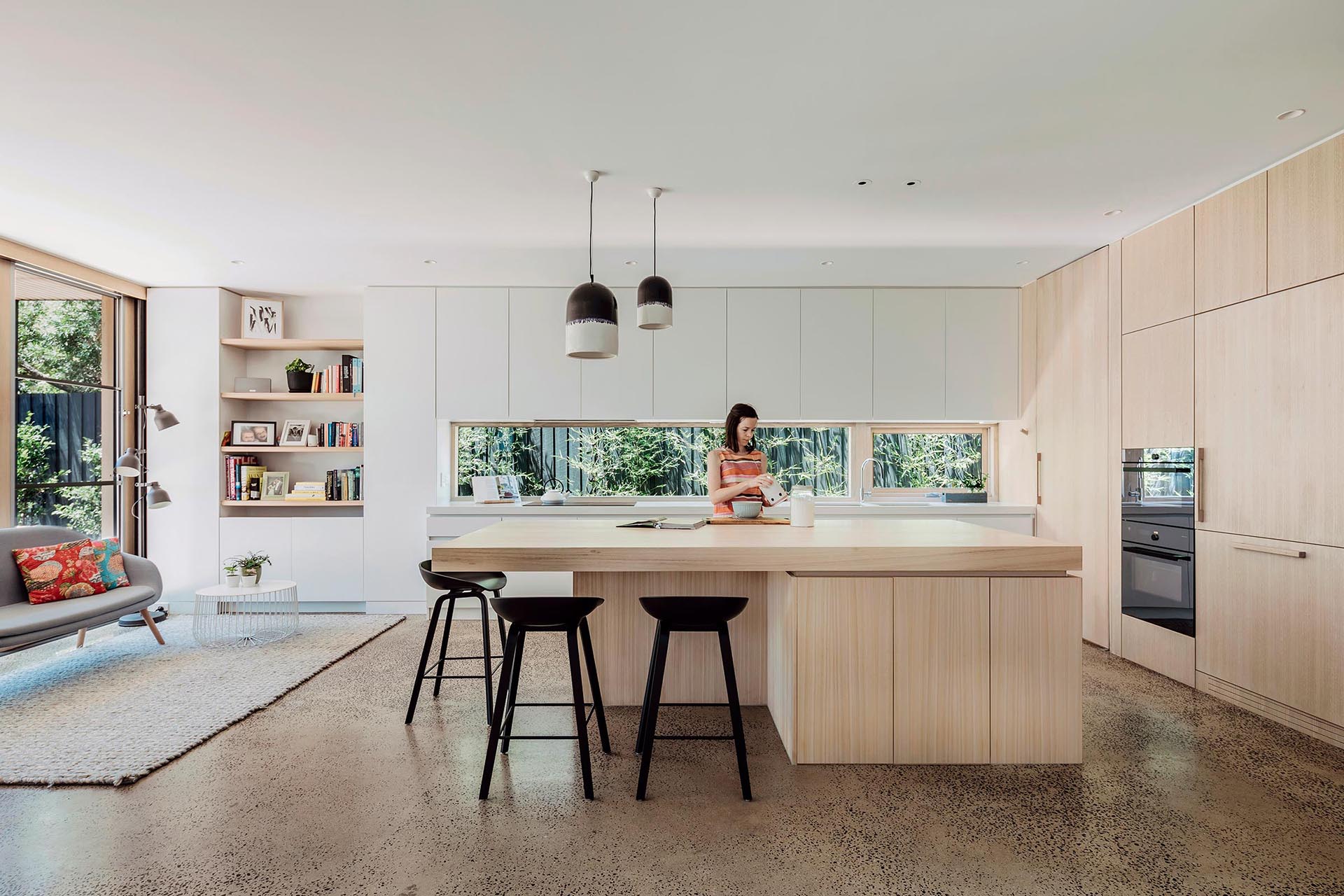 A modern open plan kitchen with minimalist white and wood cabinets, and a window that acts as a backsplash.