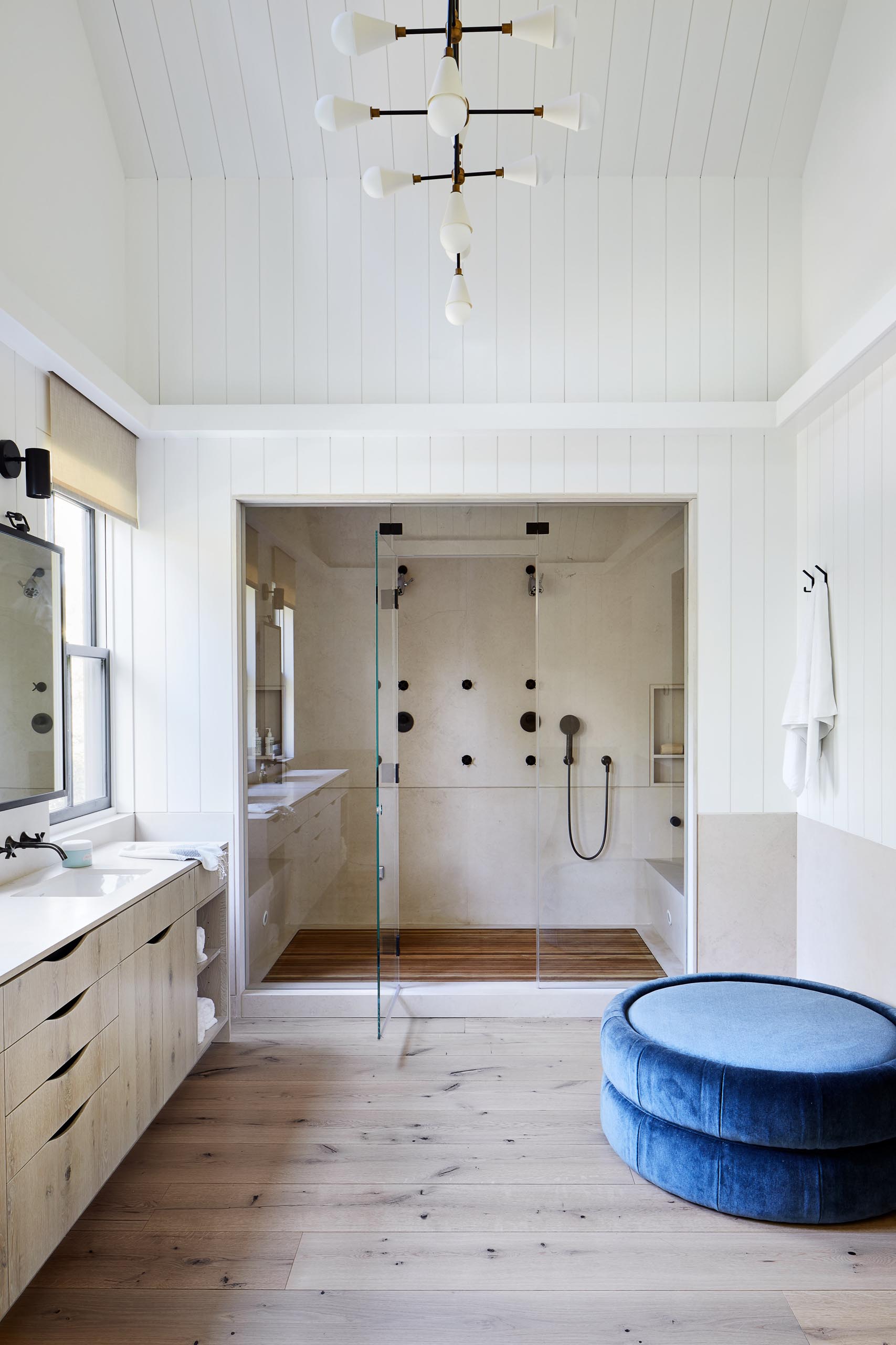 This master bathroom includes a long double sink vanity with the mirrors perfectly positioned between the black framed windows. Adjacent to the vanity is a walk-in shower with benches and double shower heads.