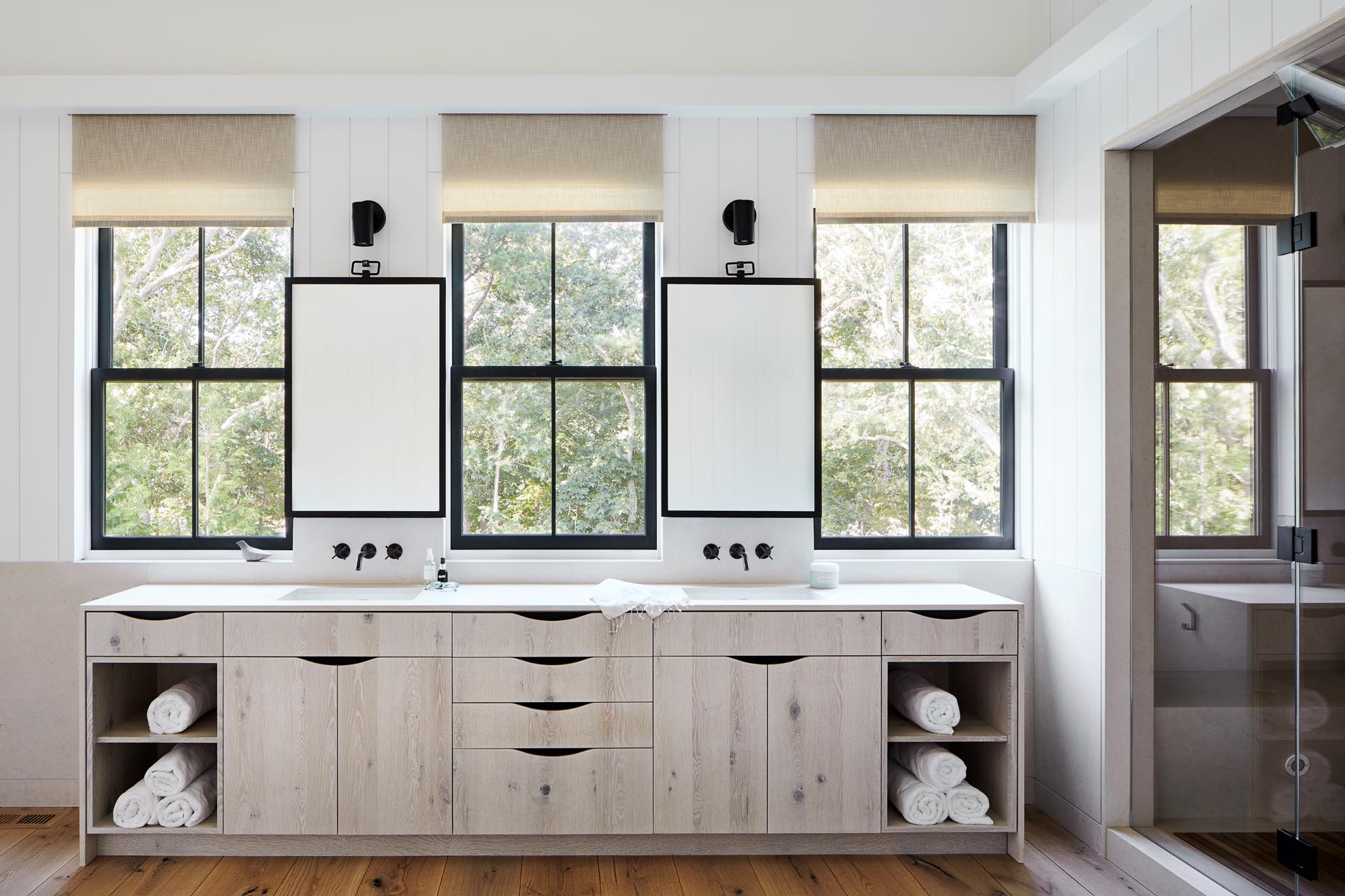 This master bathroom includes a long double sink vanity with the mirrors perfectly positioned between the black framed windows. Adjacent to the vanity is a walk-in shower with benches and double shower heads.