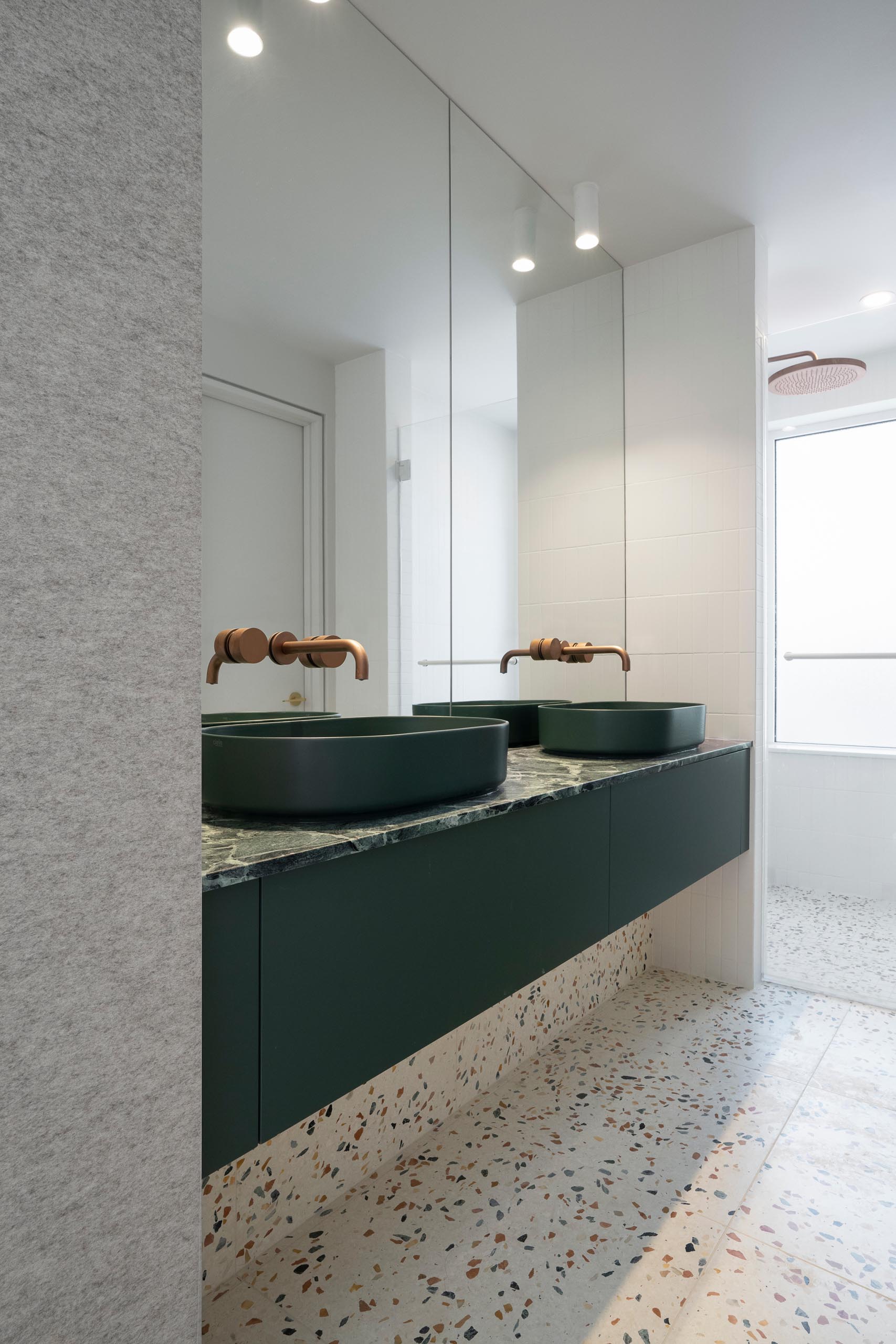 In this modern master bathroom, the Verde Saint-Denis marble countertop, emerald green sinks, and warm natural felt walls also harmonize with the terrazzo flooring.