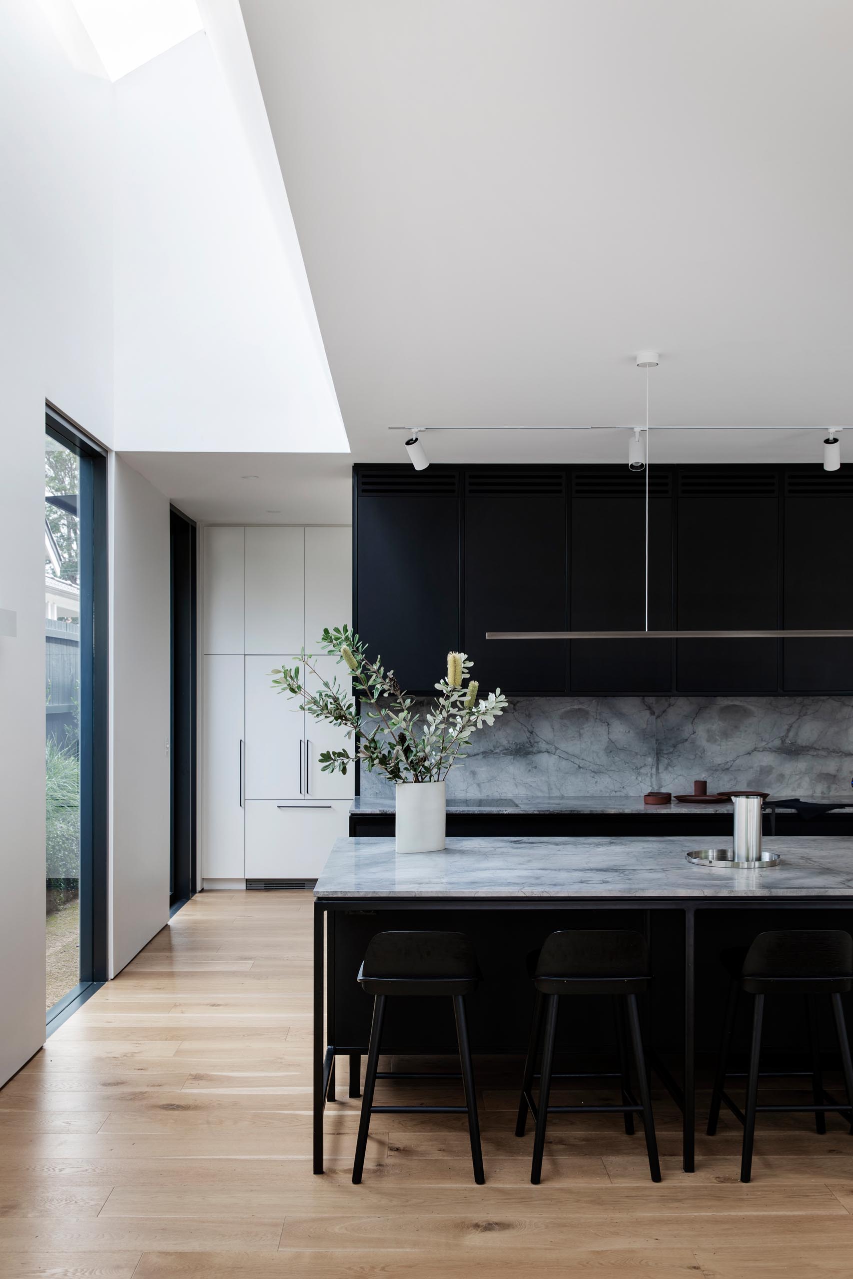 A modern kitchen has a simple color palette with matte black cabinets that are made from Paperock, a sustainable building material combining condensed layers of renewable paper bonded with resin. Grey marble, black hardware, and a minimalist horizontal light fixture complete the look.