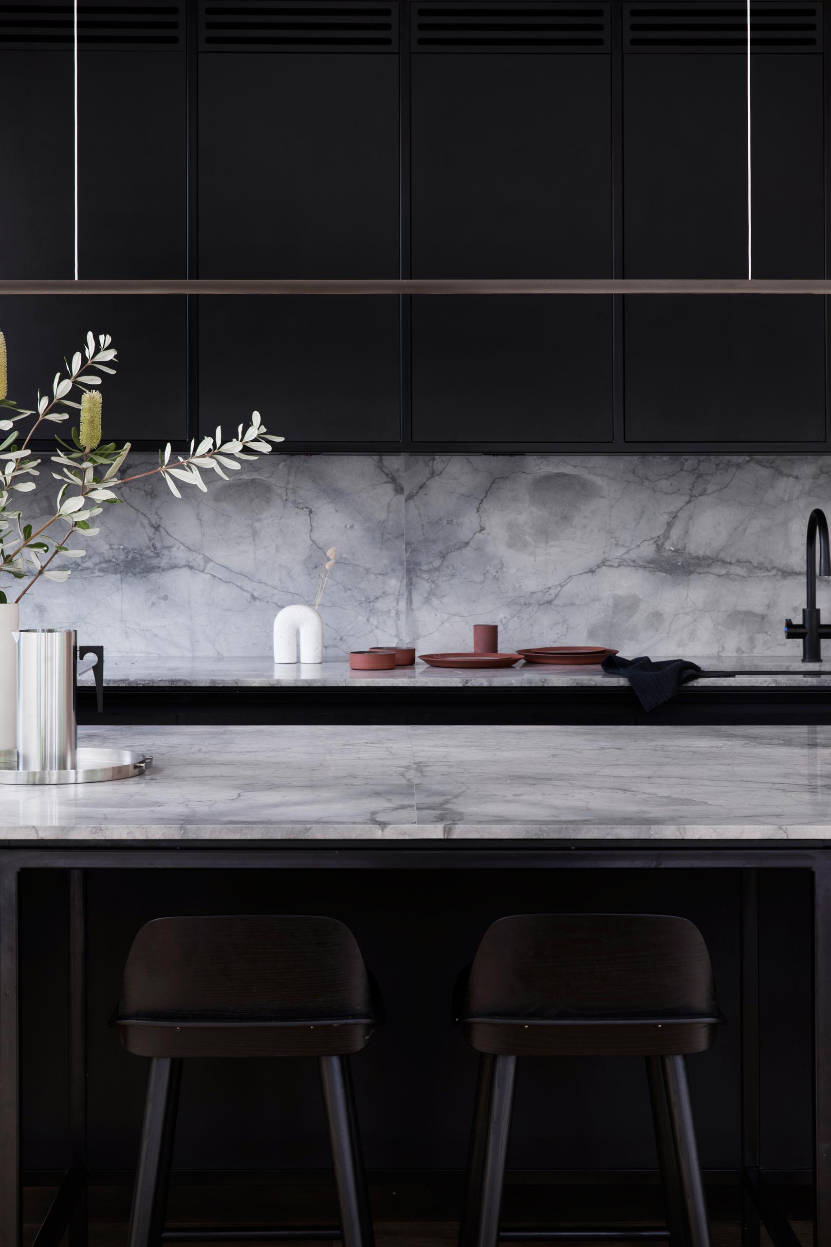 A modern kitchen has a simple color palette with matte black cabinets that are made from Paperock, a sustainable building material combining condensed layers of renewable paper bonded with resin. Grey marble, black hardware, and a minimalist horizontal light fixture complete the look.