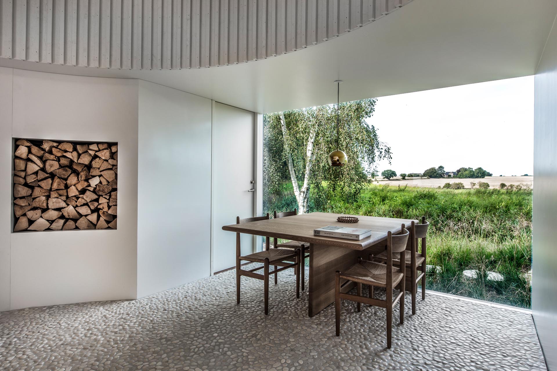 In this modern dining room, the white walls complement the painted board and batten wall cladding, which are included to provide a calm backdrop for the constantly changing textures and colors of the agricultural landscape seen through the window.