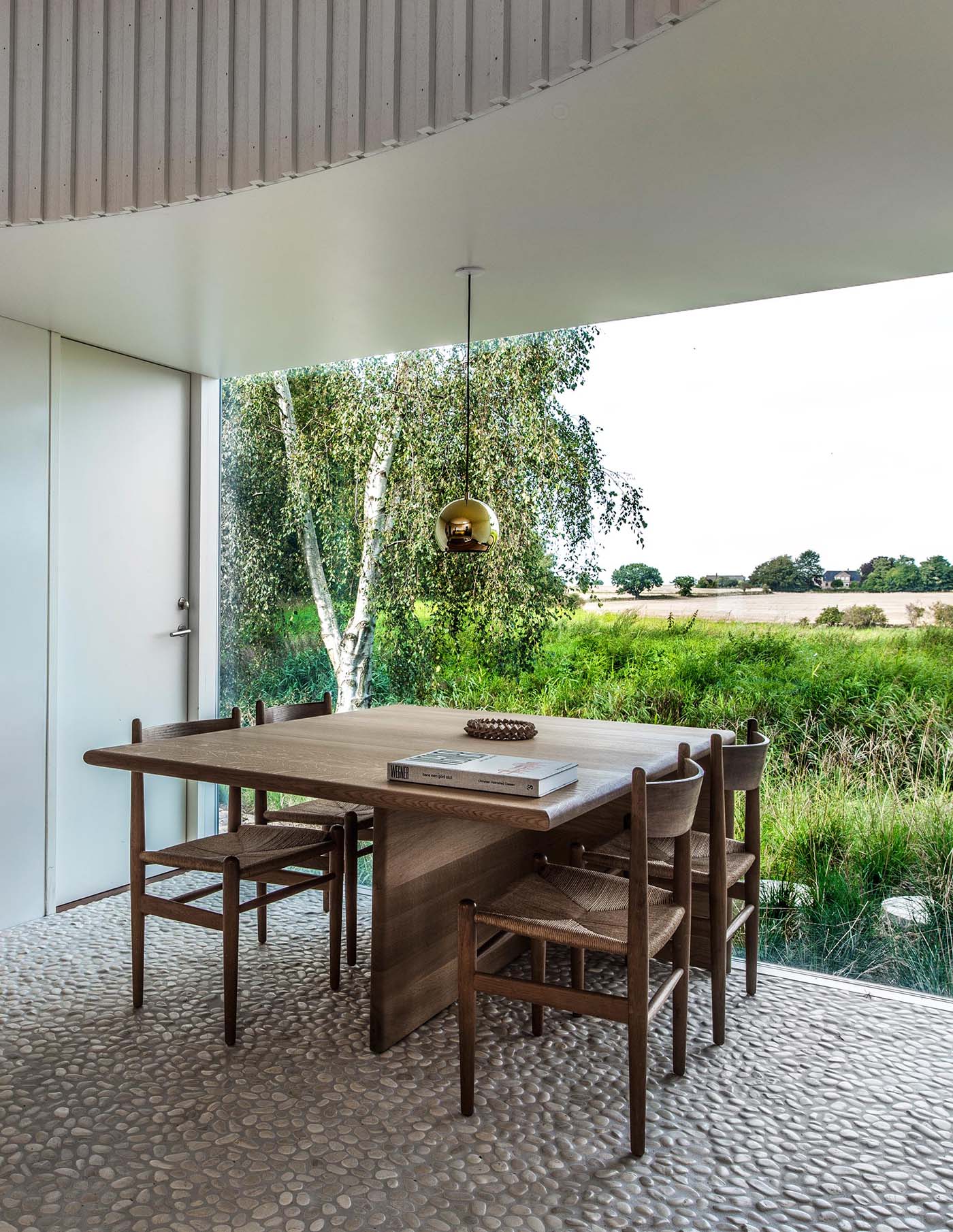 In this modern dining room, the white walls complement the painted board and batten wall cladding, which are included to provide a calm backdrop for the constantly changing textures and colors of the agricultural landscape seen through the window.