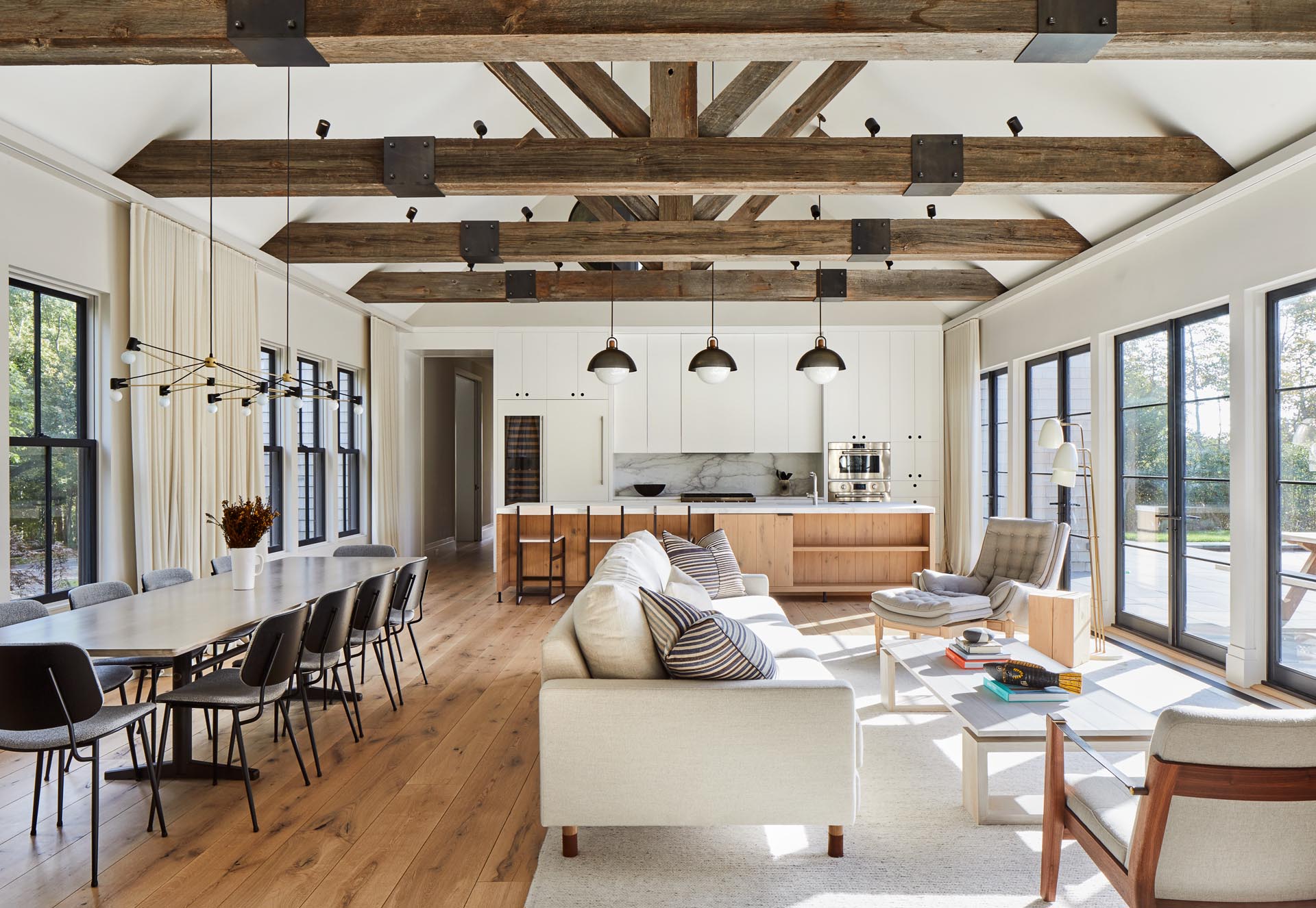 This modern farmhouse great room has plenty of room for the living room, dining area, and kitchen, with exposed beams, and windows on one side, and doors on the other.