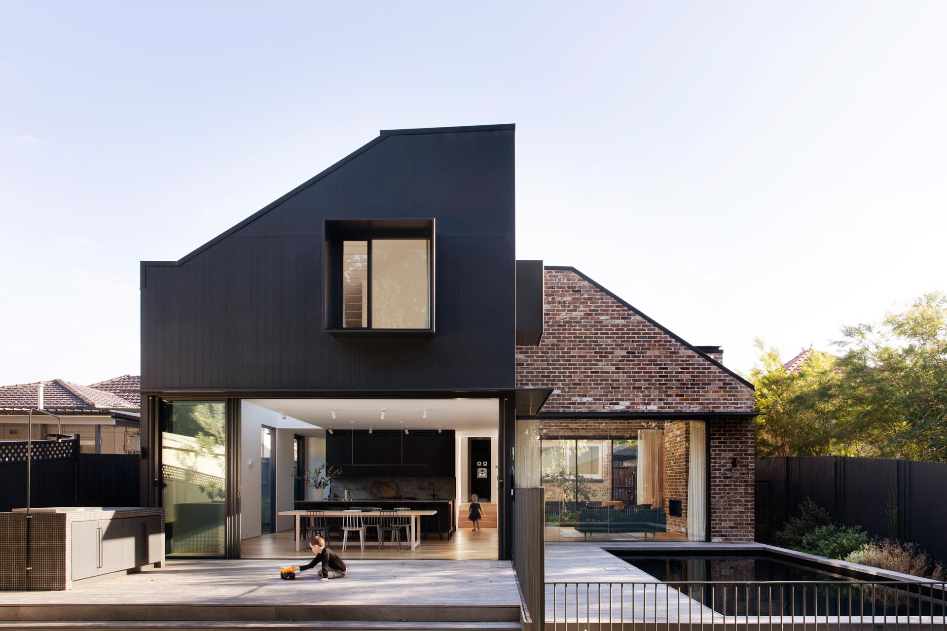 A sliding glass wall opens the interior spaces of a modern addition to a greyed Blackbutt deck with an outdoor kitchen that steps down to the yard, as well as a swimming pool.