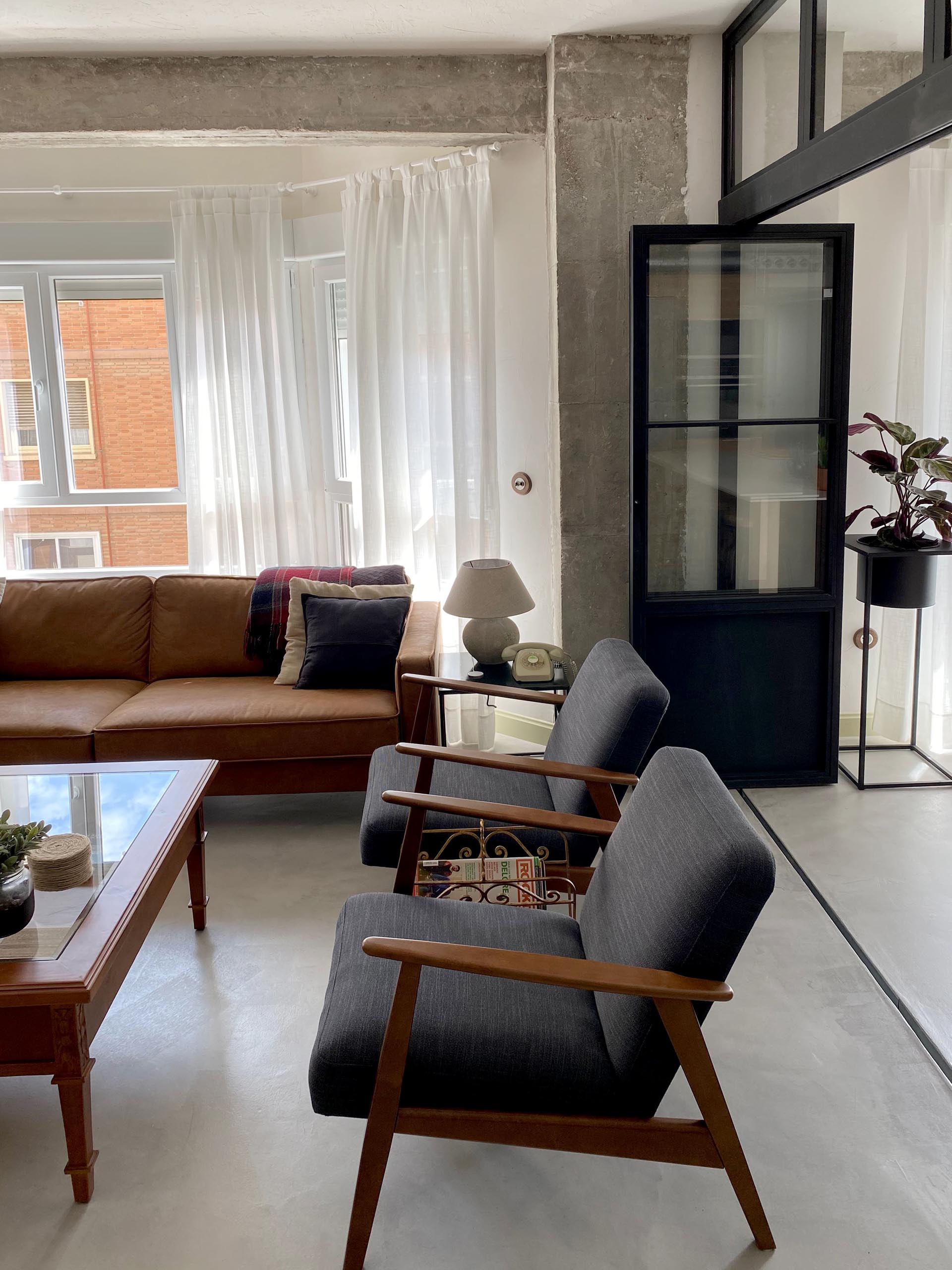 This modern living room showcases the concrete flooring, and is furnished with leather couches, a pair of armchairs, and a wood coffee table. On the living room wall, there's a low console and glass front cabinet that complements the black framed doors.