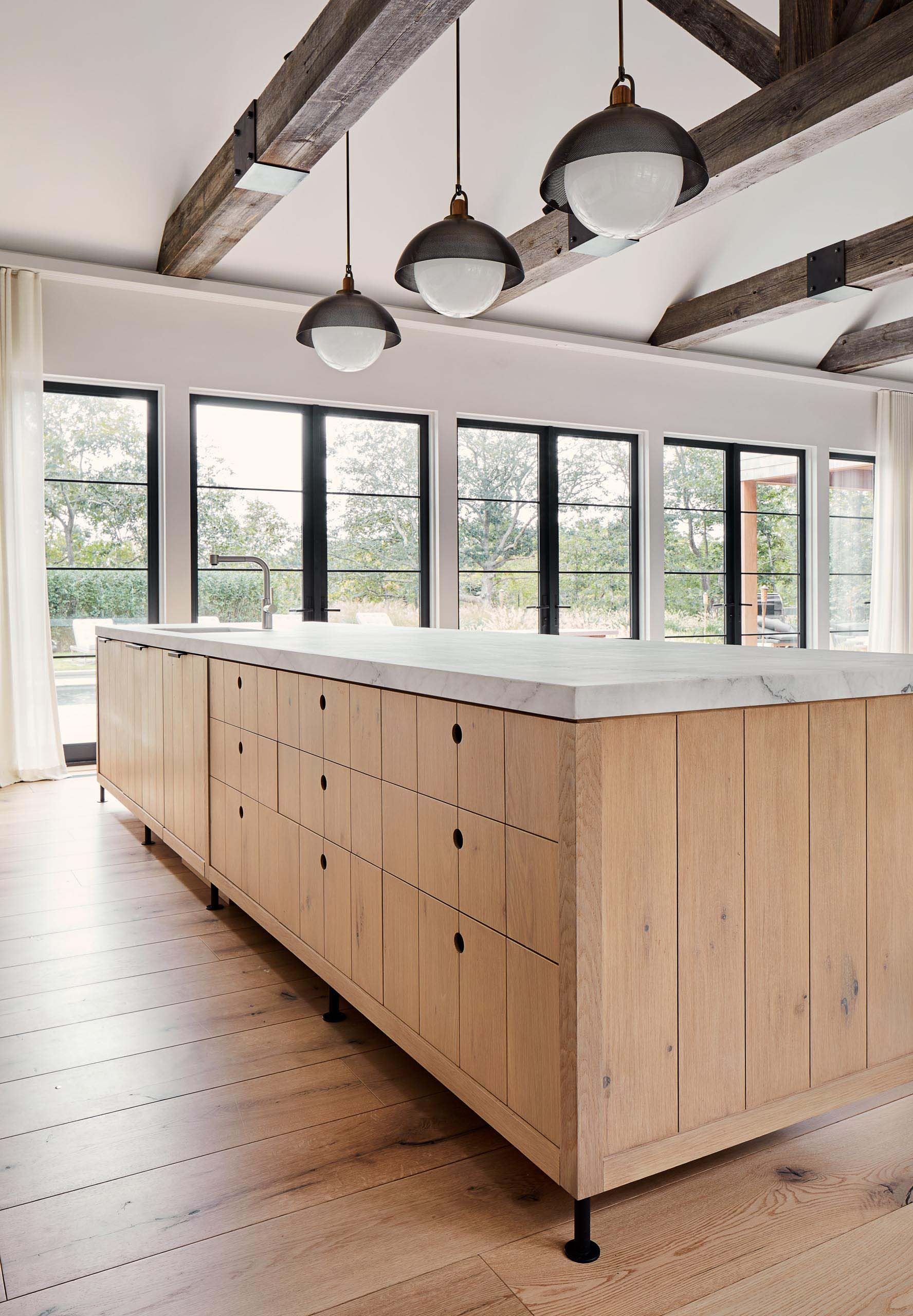 This modern farmhouse kitchen island has wood cabinets and drawers, while hanging above the island are pendant lights from Allied Maker, and the stools are from Skram Furniture Co.