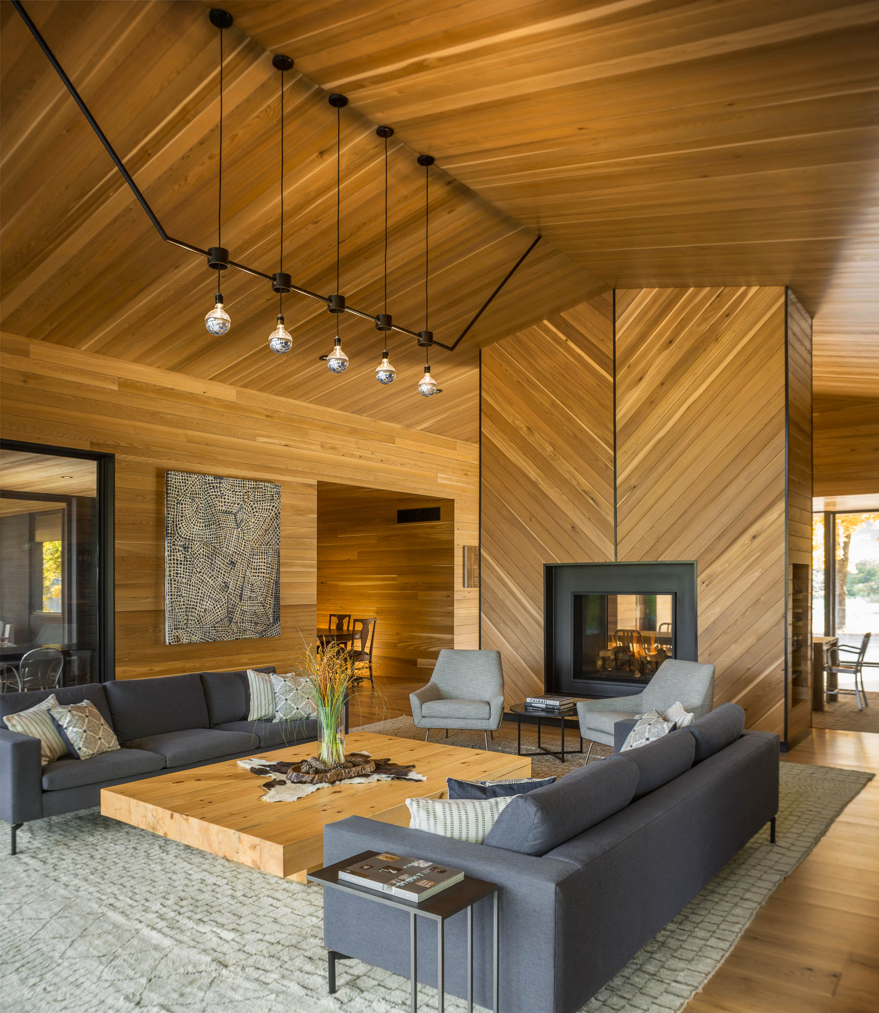 A modern living room with a glass front fireplace, a large coffee wood coffee table, a unique lighting element, and wood-lined walls and ceiling.