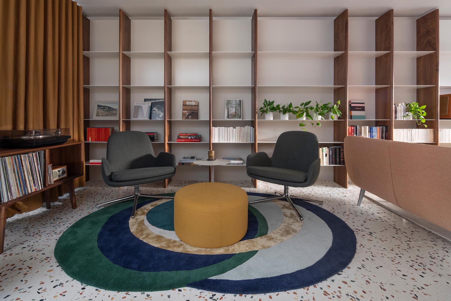 This modern living room with a terrazzo tiled floor, receives plenty of natural light, while a custom wood and metal shelving unit covers the entire wall and expands through to a secondary seating area with a colorful rug and record player.